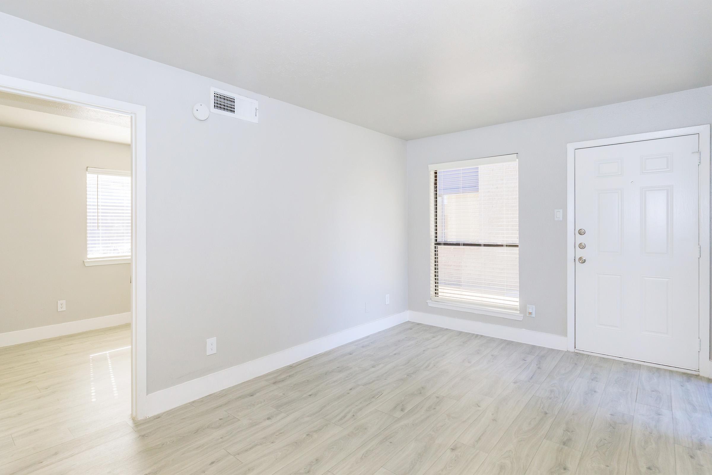 A brightly lit, empty living space with light gray walls and wood-style flooring. One corner features a window with blinds, while another has a white door leading outside. A small doorway on the left suggests access to another room. The overall atmosphere is open and airy.