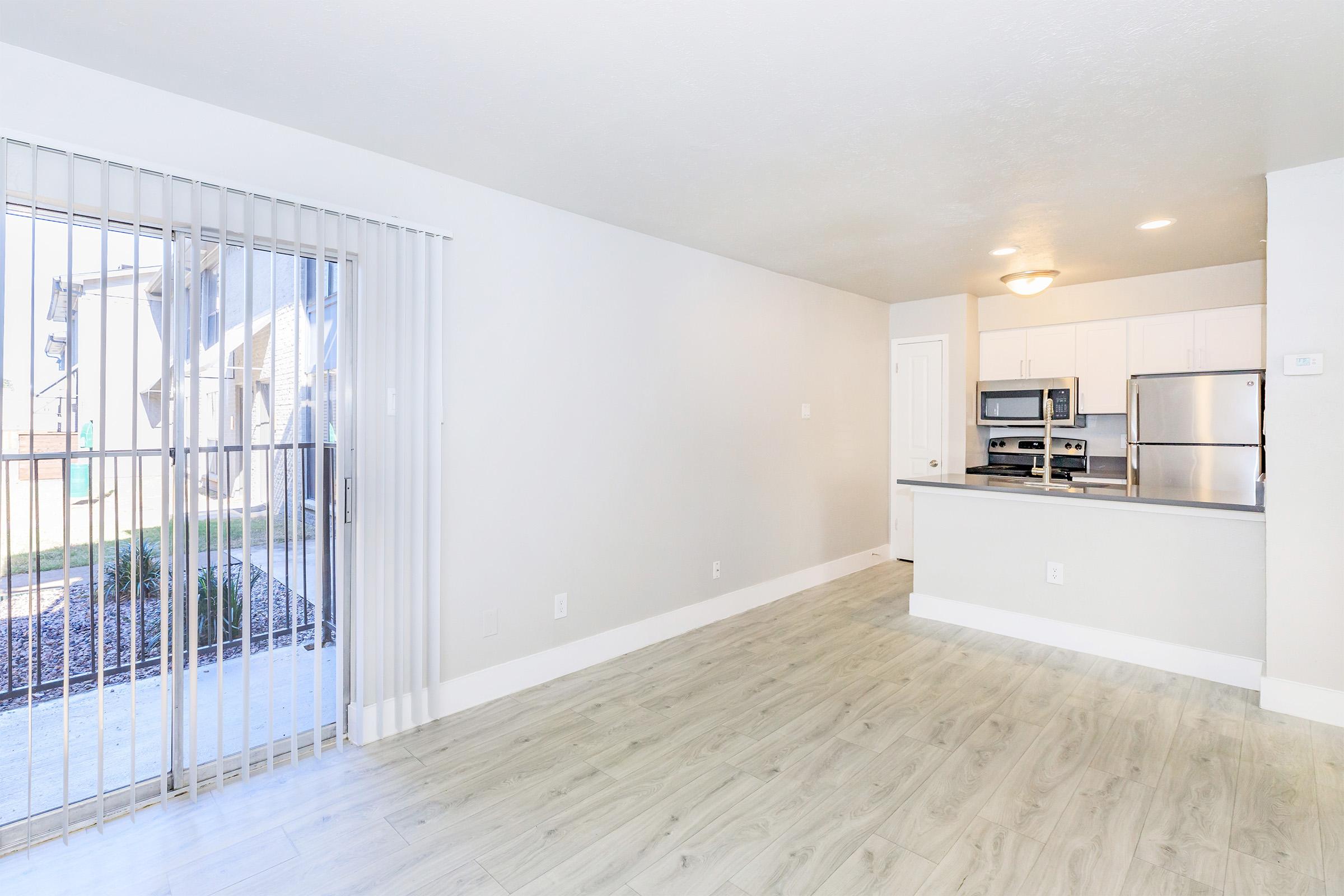 Bright, open living space featuring light-colored walls and modern wood flooring. A sliding glass door leads to an outdoor area, while a contemporary kitchen with stainless steel appliances and a breakfast bar is visible in the background. Ideal for a cozy yet stylish living environment.