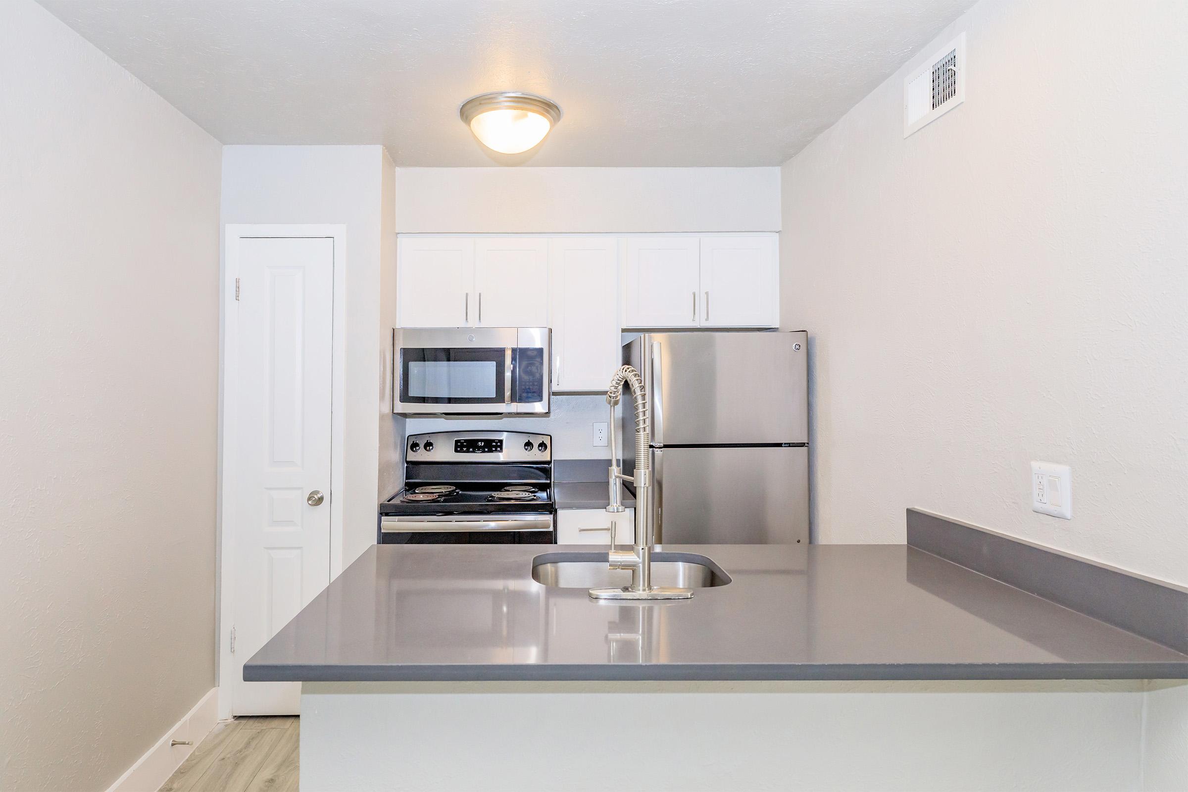 Modern kitchen featuring gray countertops, stainless steel appliances including a refrigerator and microwave, white cabinets, and a sink with a pull-down faucet. The walls are painted light gray, contributing to a bright and airy atmosphere. A door to the left leads to another room.
