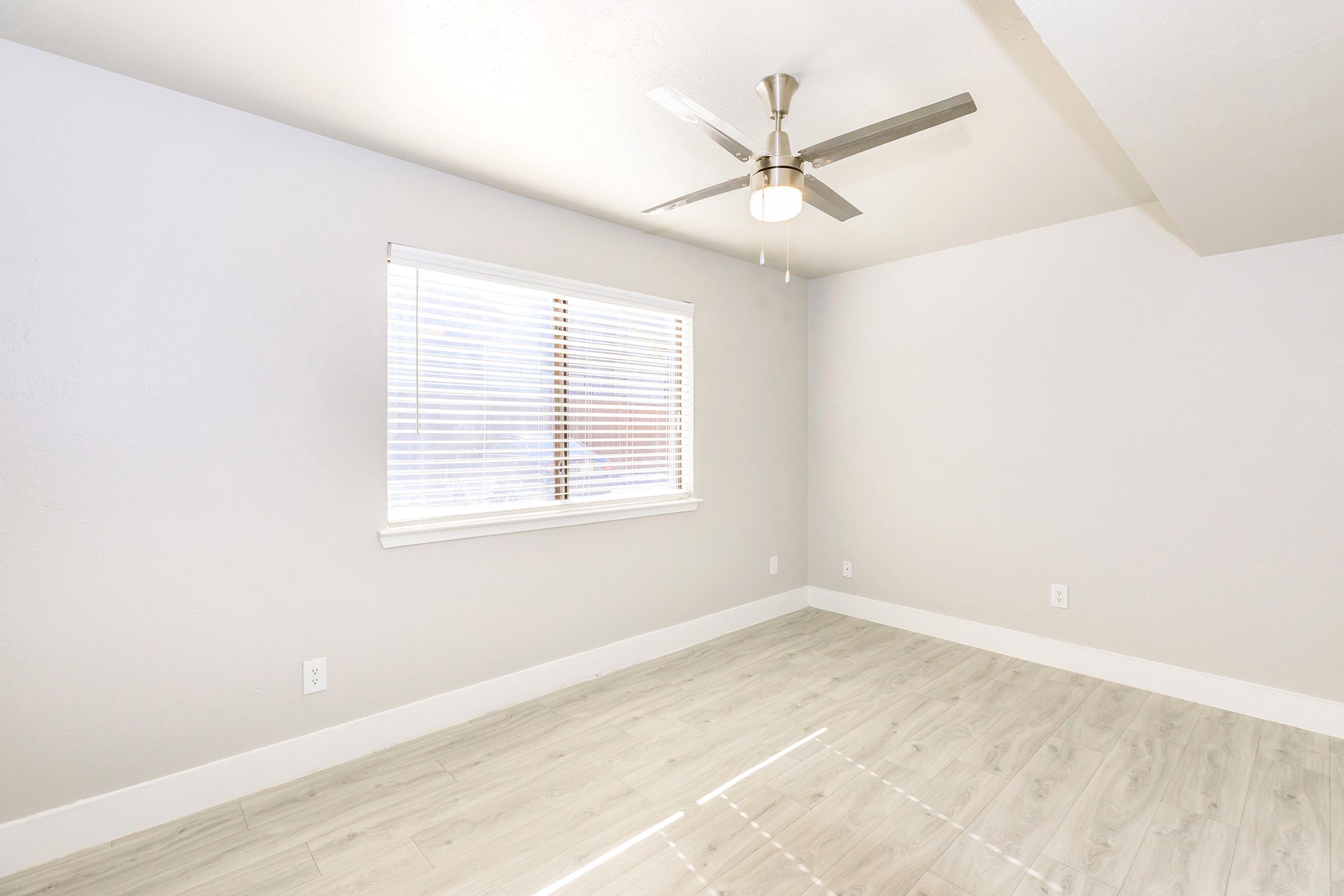 A clean, empty room with light gray walls and a light-colored wooden floor. The room features a ceiling fan and a window with white blinds, allowing natural light to enter. The space appears bright and modern, with minimalistic decor.