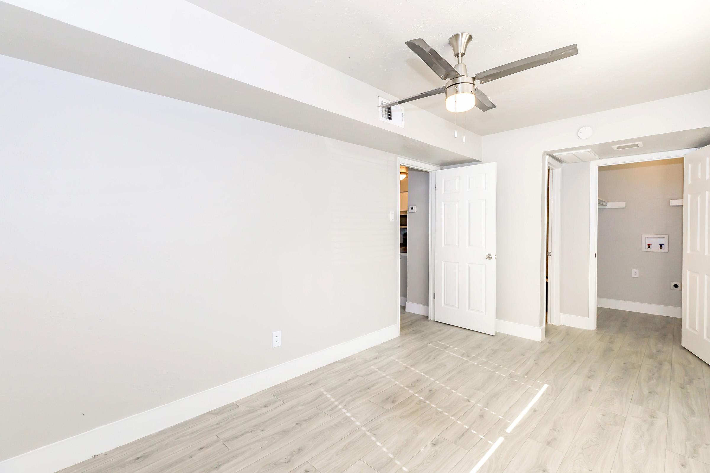 A bright, modern room featuring light gray walls and light wood flooring. It has a ceiling fan with a central light fixture. On one side, there are double doors leading to another space, while the opposite side has a single door. Natural light streams in, creating an inviting atmosphere.