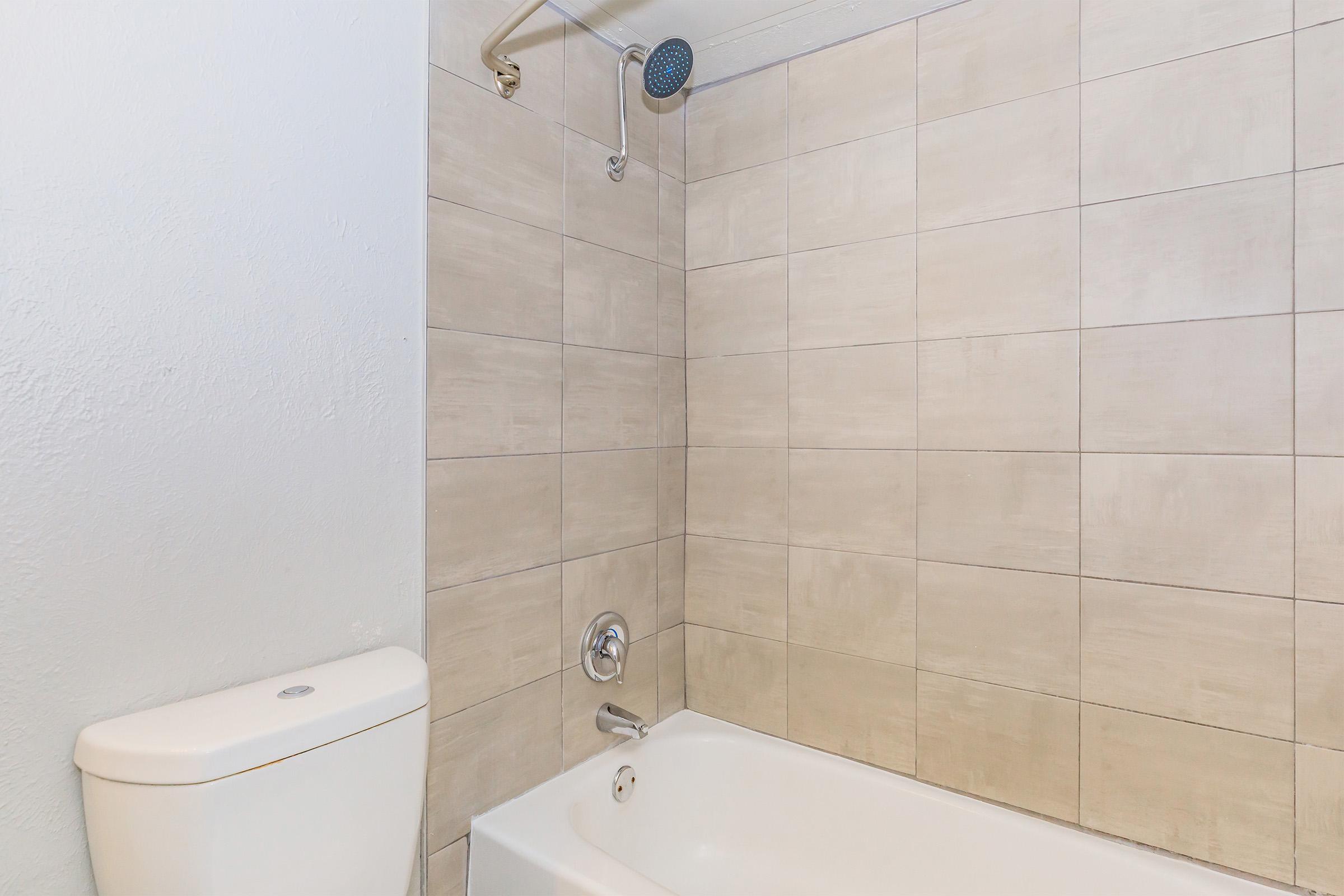 A clean bathroom featuring a bathtub with a showerhead, beige tiled walls, and a white toilet. The space is well-lit, highlighting the simple and modern design.