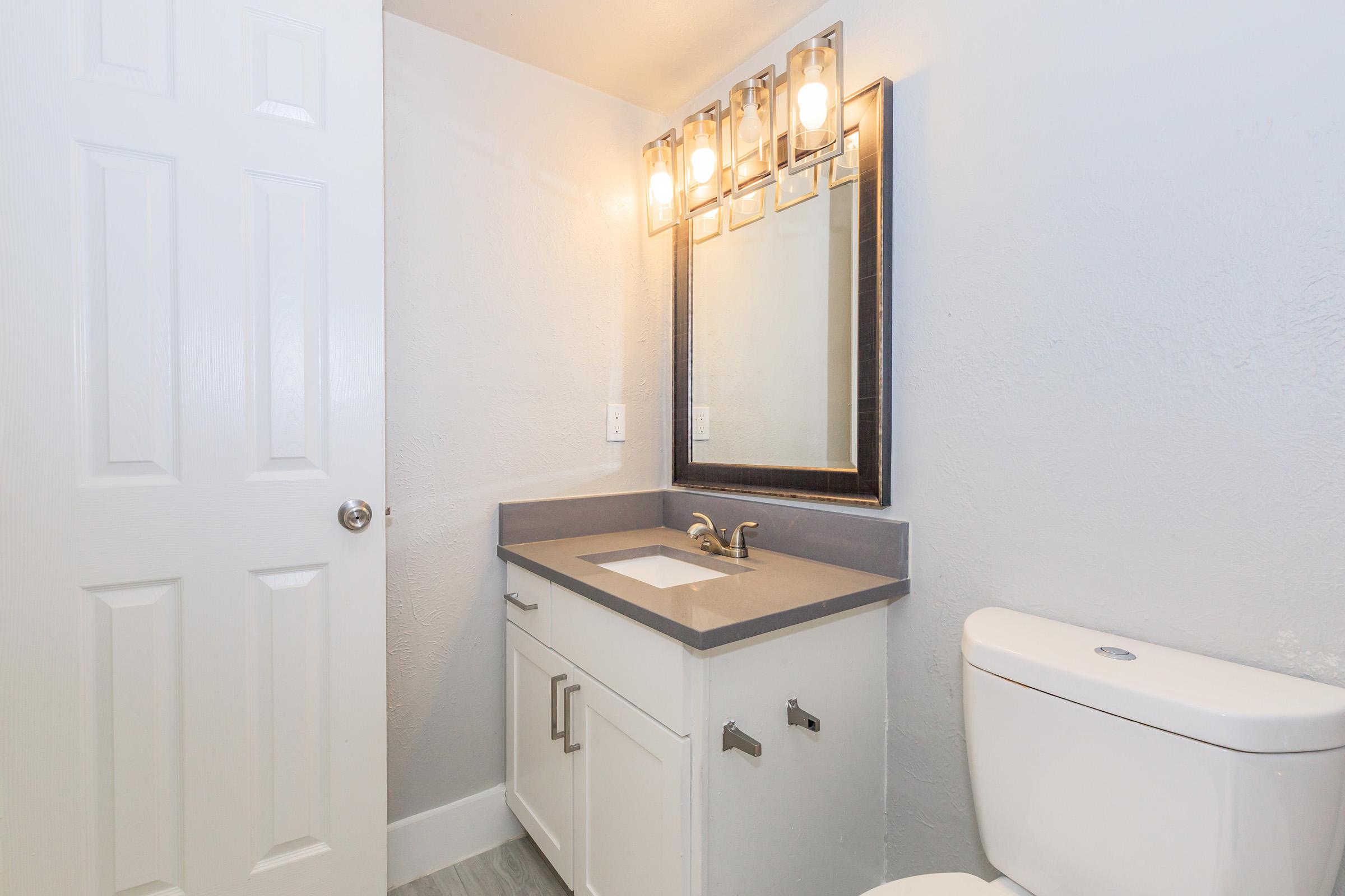 A modern bathroom featuring a gray countertop with a sink, a large illuminated mirror above it, and a white toilet. The walls are a light color, and there is a white door on one side. The overall design is clean and contemporary.