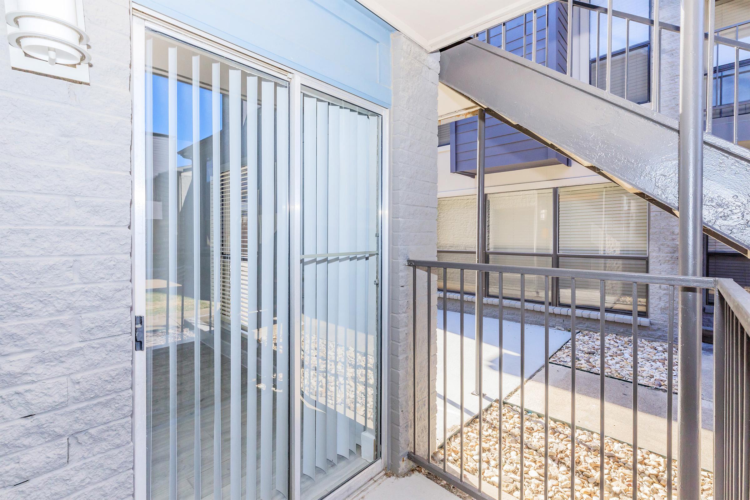 A balcony view of an apartment entrance featuring a sliding glass door with vertical blinds, a metal railing, and a staircase. The area includes light-colored stone pebbles and walls, with natural light illuminating the space. Windows are visible in the background.