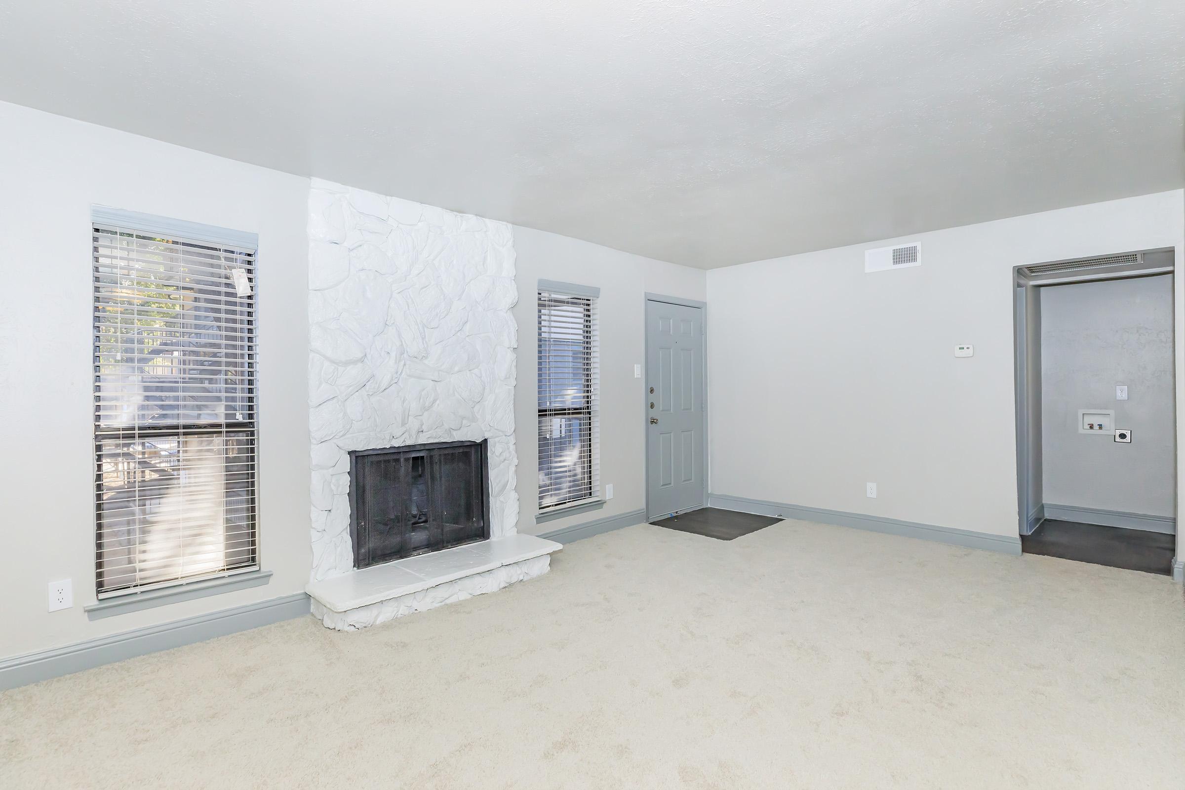 A spacious living room with light gray walls, a white stone fireplace, and large windows featuring blinds. The floor is covered in a light beige carpet. To the right, there is a gray door leading to another room or hallway. Overall, the space is bright and inviting.