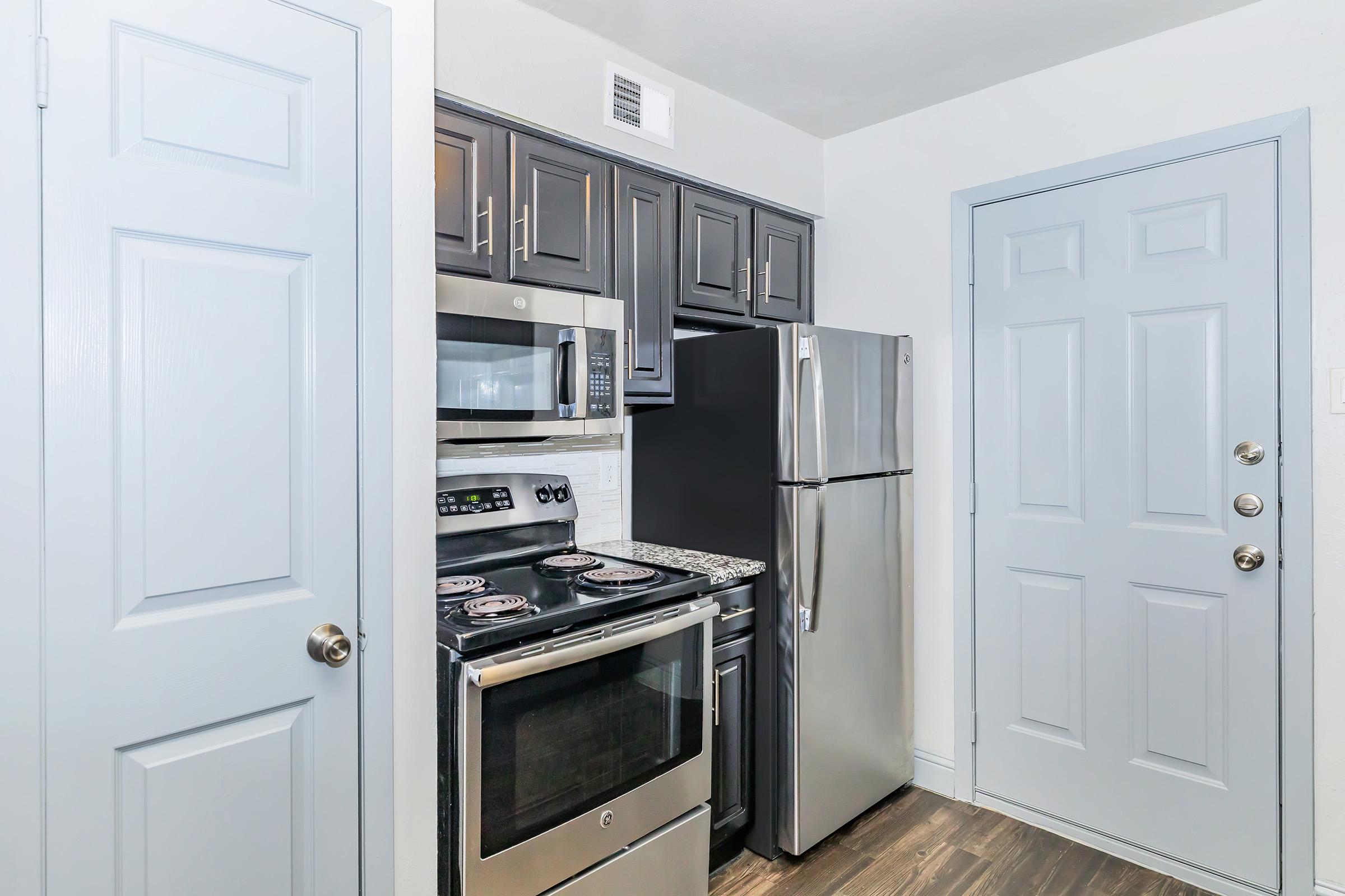 A modern kitchen featuring dark cabinetry, stainless steel appliances including a stove and refrigerator, and gray walls. A door leading outside is visible, along with a light-colored door. The space is well-lit and designed for functionality.