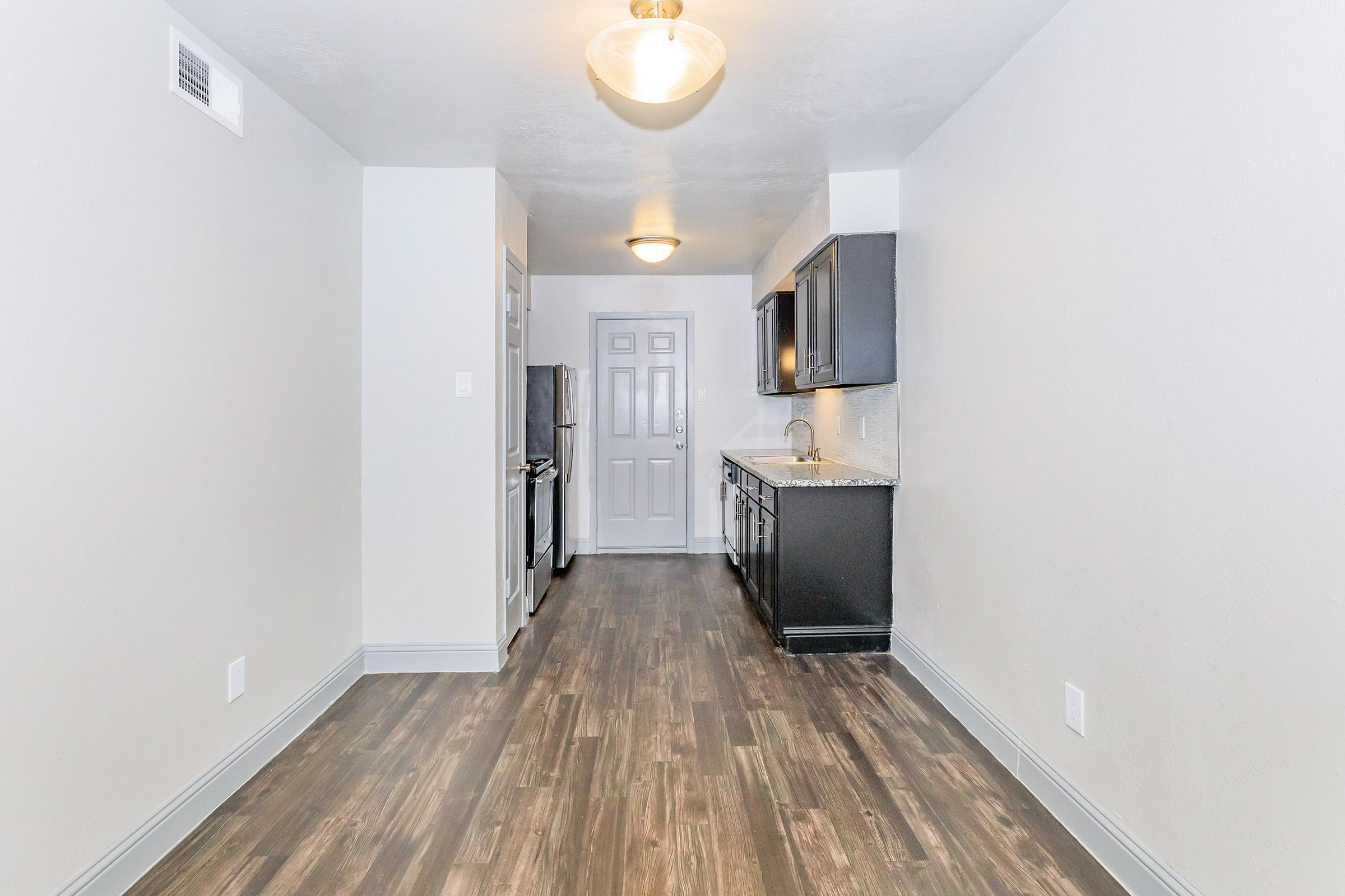 A modern kitchen featuring dark cabinets, granite countertops, and stainless steel appliances. The space has a light-colored wall and a wood-like floor, with a light fixture hanging from the ceiling. There's a doorway leading to another room at the far end.