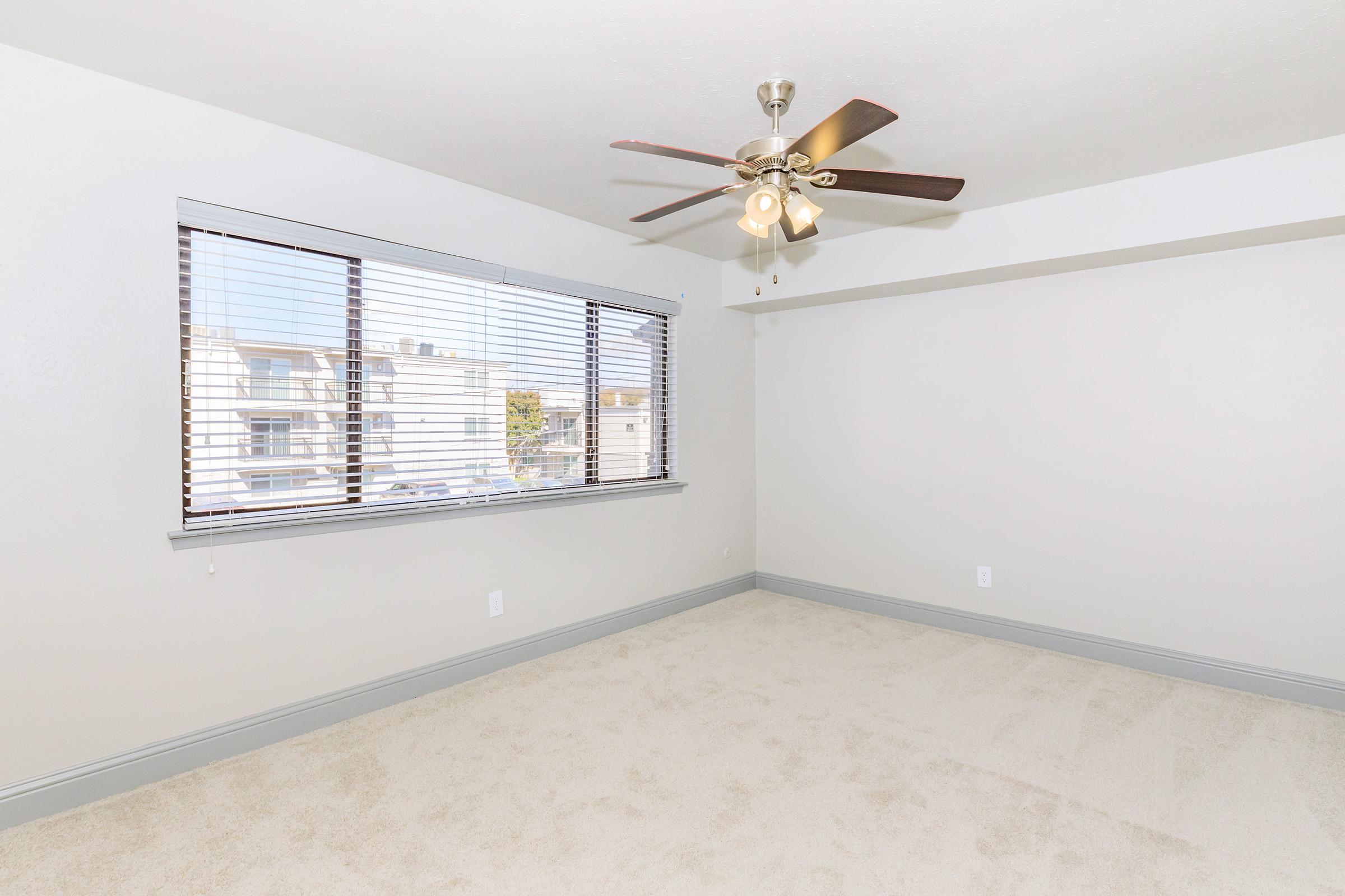 A spacious, empty room featuring light beige carpet, a ceiling fan with five blades, and large windows with wooden blinds. The walls are painted in a light shade, and there's a baseboard along the floor. Natural light fills the space, creating a bright and airy atmosphere.
