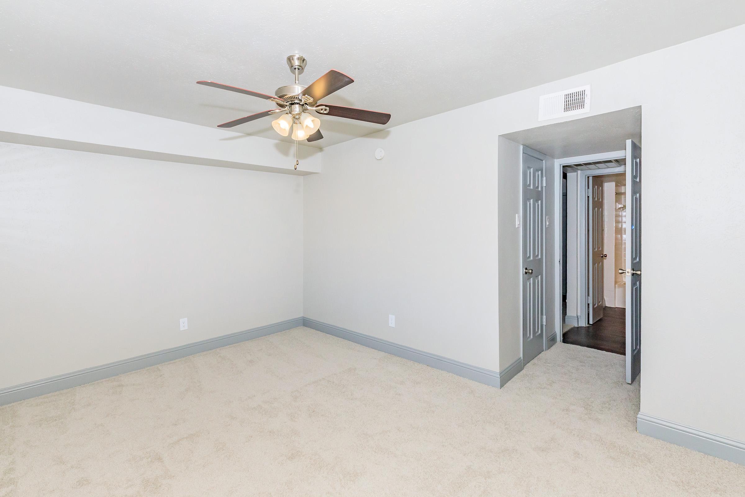 A spacious room with light-colored walls and a ceiling fan. The floor is covered in beige carpet, and there is a doorway leading to another room. Natural light may enter from the adjacent area, enhancing the airy feel of the space.