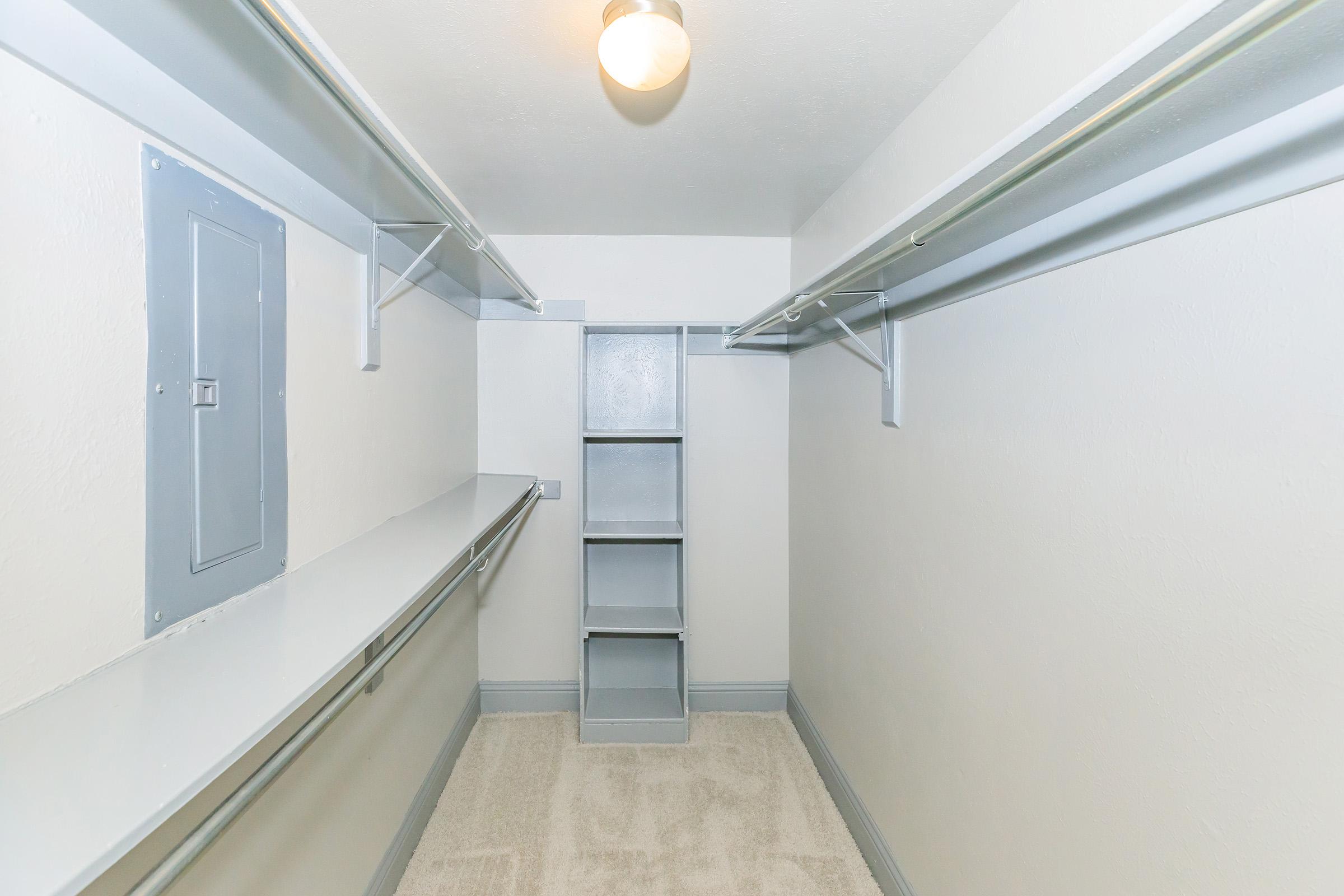 A spacious, empty walk-in closet featuring light gray walls and shelving on both sides. There is a built-in shelf unit in the center, a ceiling light fixture, and a clean carpeted floor, creating a bright and organized storage space.