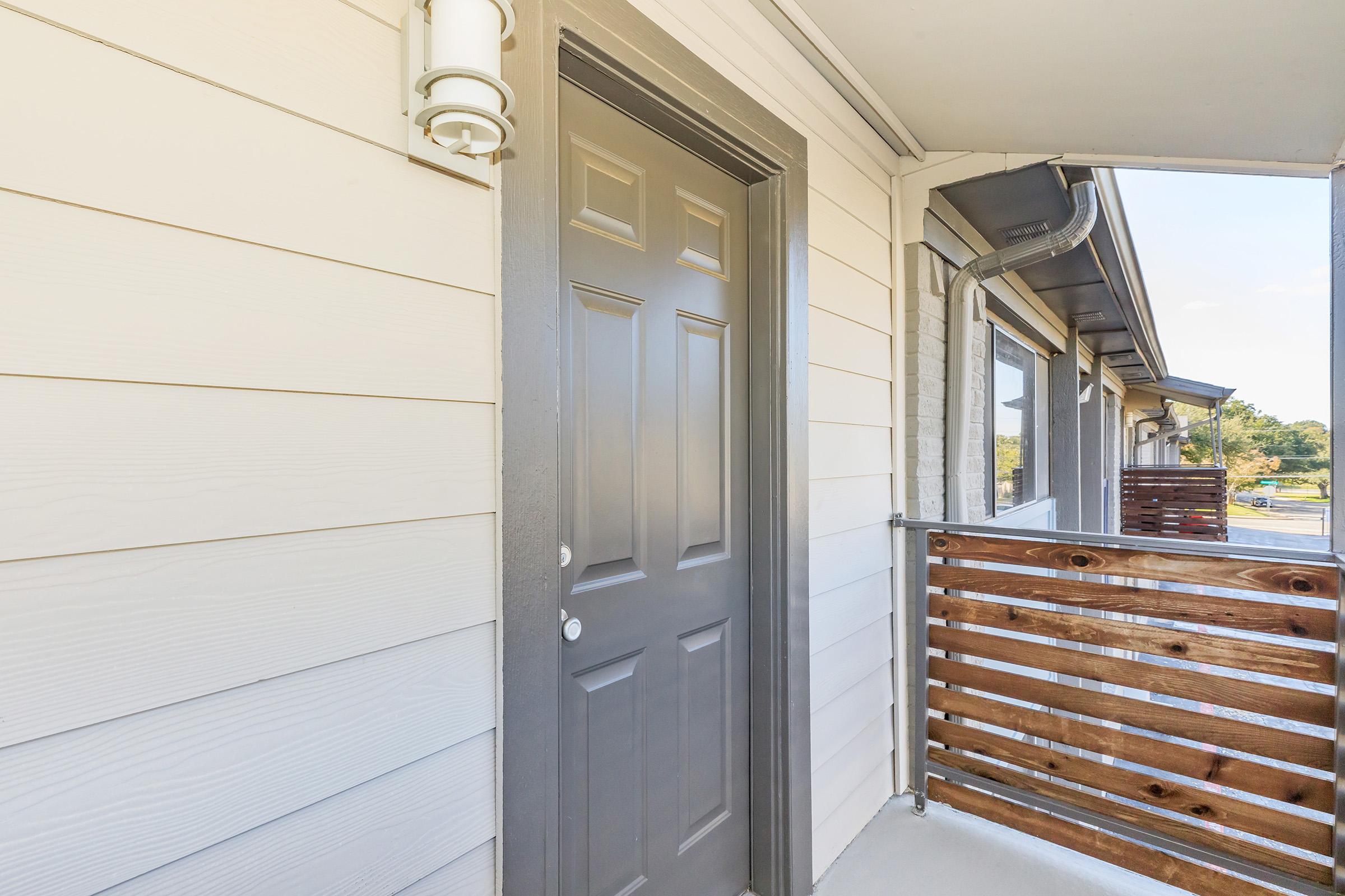 A close-up view of a light-colored exterior wall featuring a dark gray door. The door has a simple design with a round doorknob and a vertical panel. There is a light fixture next to the door and a wooden railing visible, indicating an outdoor balcony area. The setting appears to be a residential or apartment building.