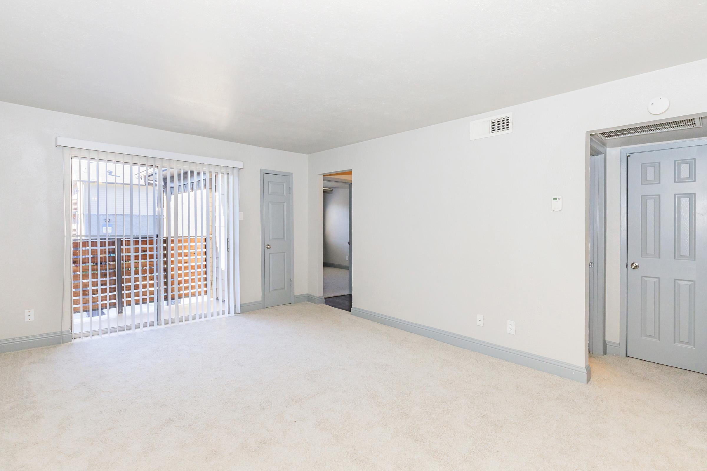 Spacious, empty room with light gray walls and cream-colored carpet. Features sliding glass doors with vertical blinds leading to a small balcony. Two doors on opposite walls suggest access to other rooms. Brightly lit with natural light coming in from the balcony area.