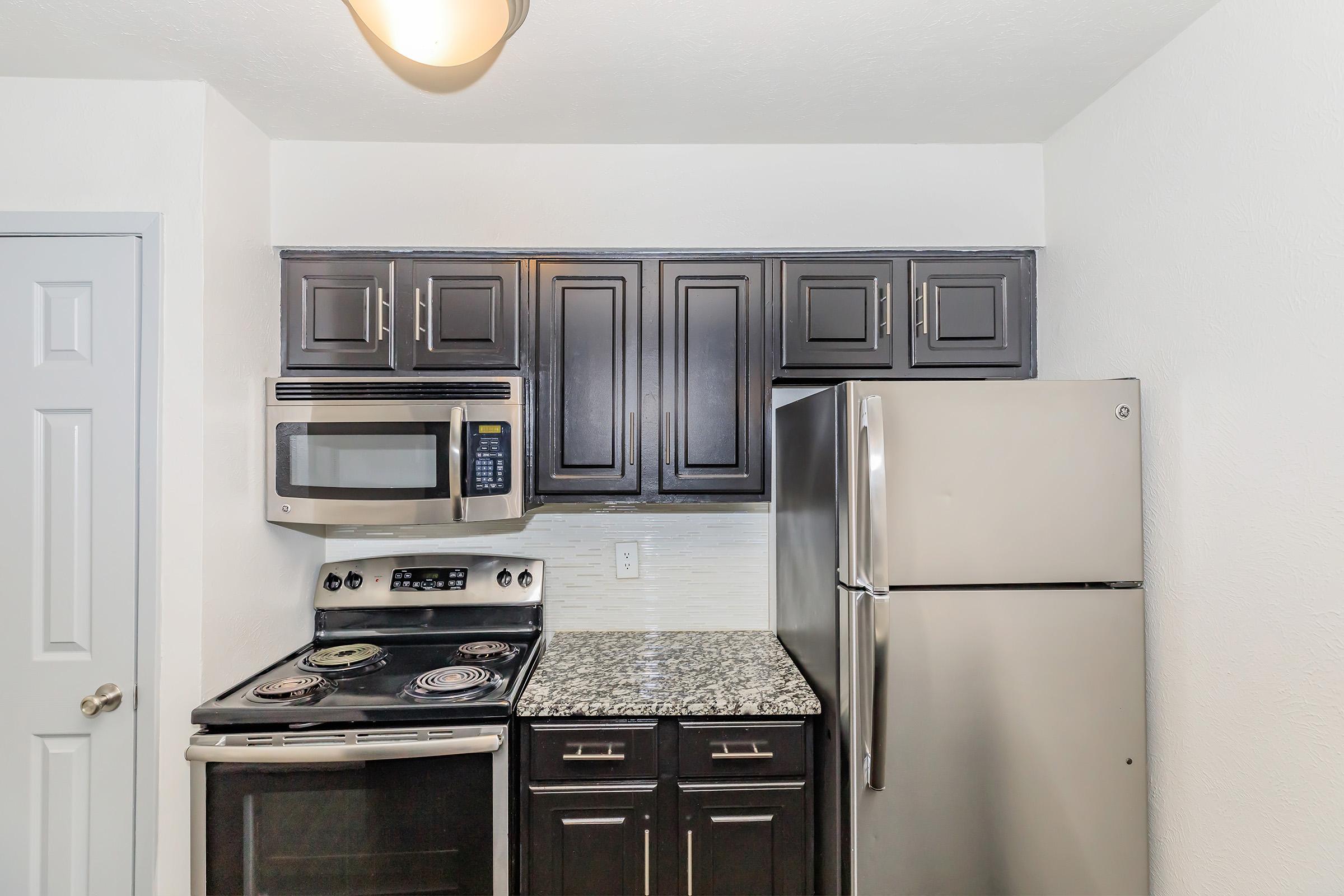 A modern kitchen featuring dark wood cabinets, a black stove, a microwave, and a stainless steel refrigerator. The countertop is made of granite, and there is a neutral-colored backsplash, creating a sleek and functional cooking space.