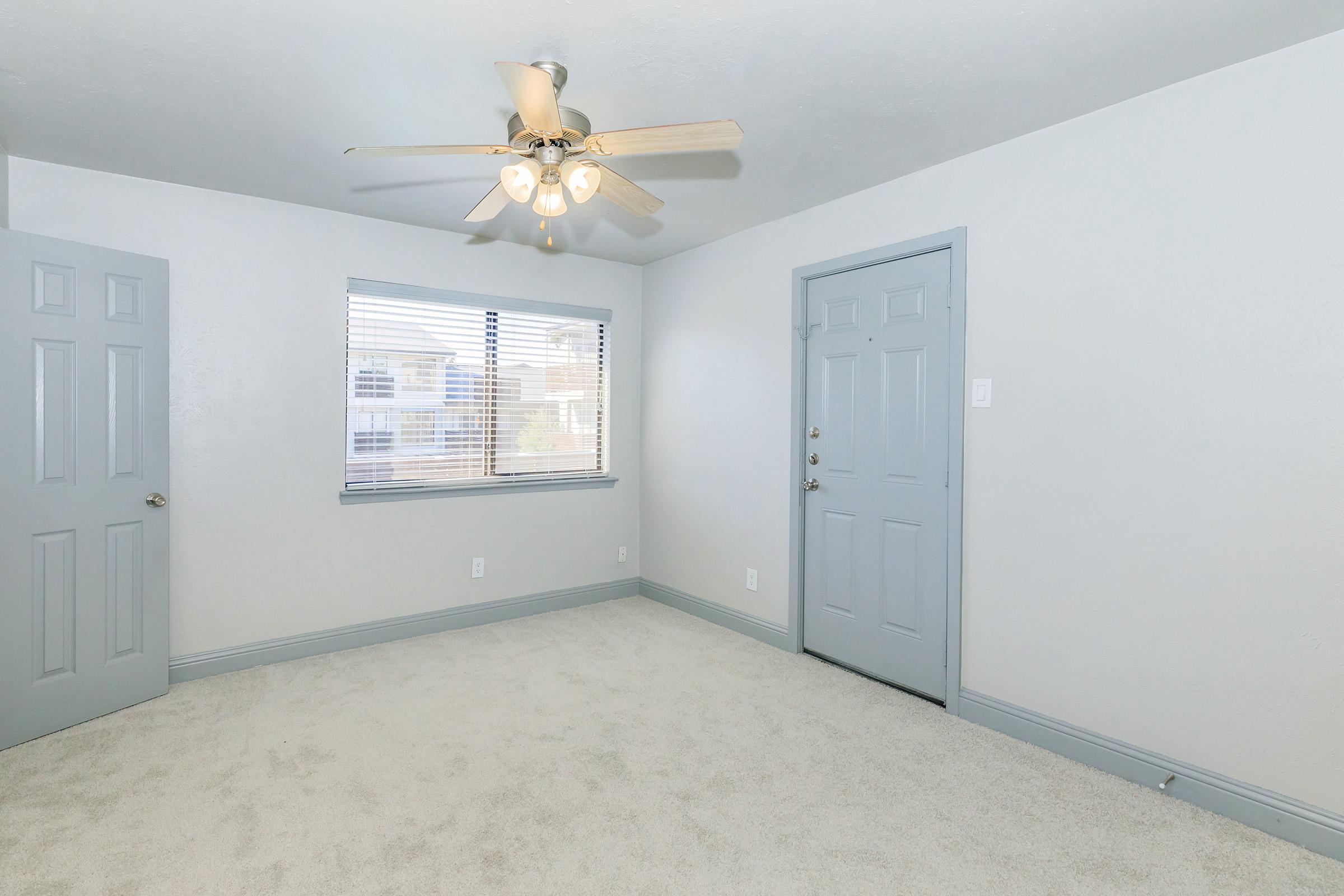 An empty room with light gray walls and a ceiling fan. There are two doors: one leading outside and another possibly to a closet. A window with blinds allows natural light to enter. The floor is covered with light-colored carpet, giving the space a clean and airy feel.