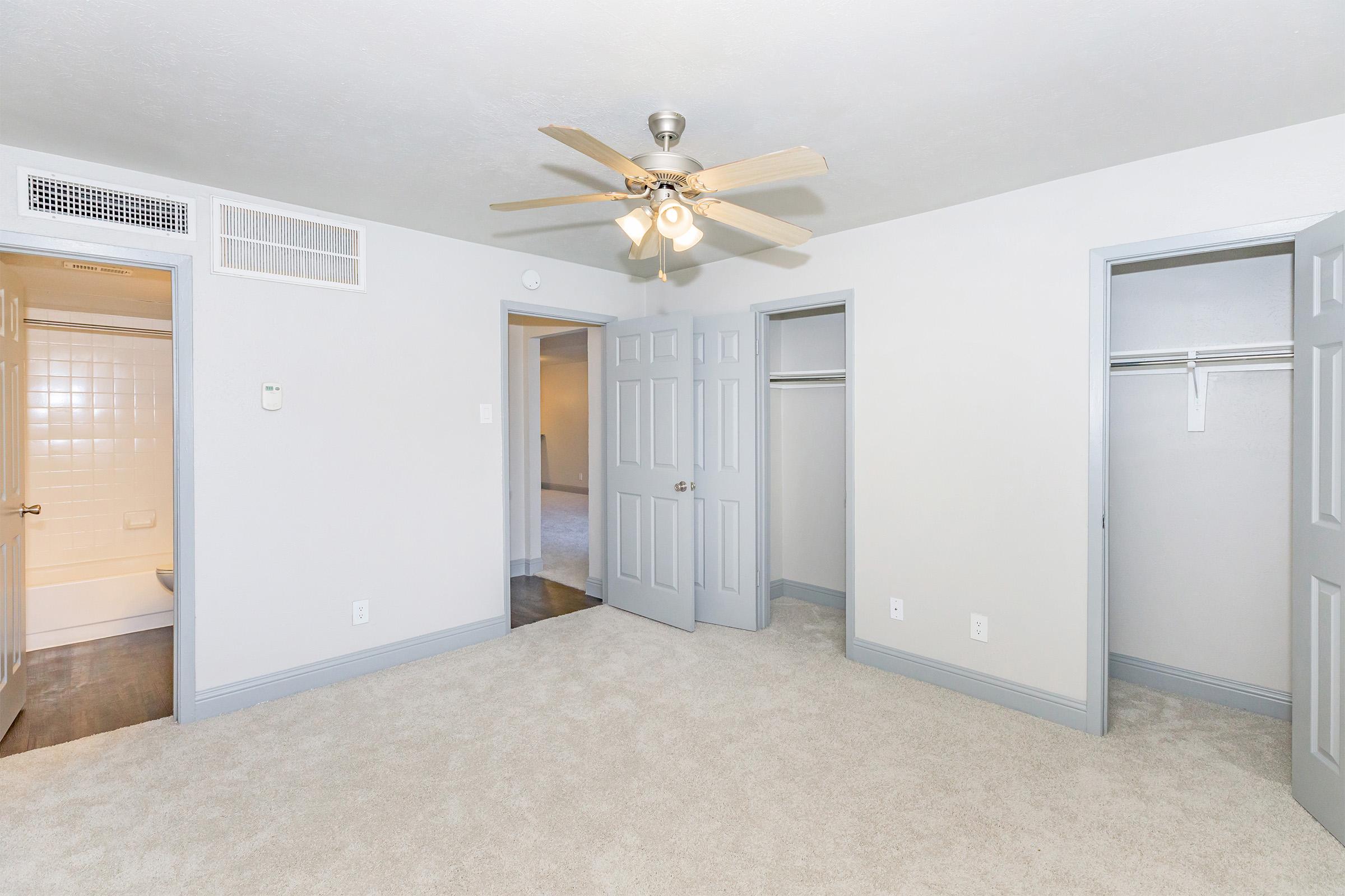 A bright, neutral-colored bedroom featuring soft carpeting and a ceiling fan. There are two doors leading to closets, each with double doors. Another door is visible, presumably leading to a bathroom. The light walls enhance the room's spacious feel, with natural light likely streaming in.