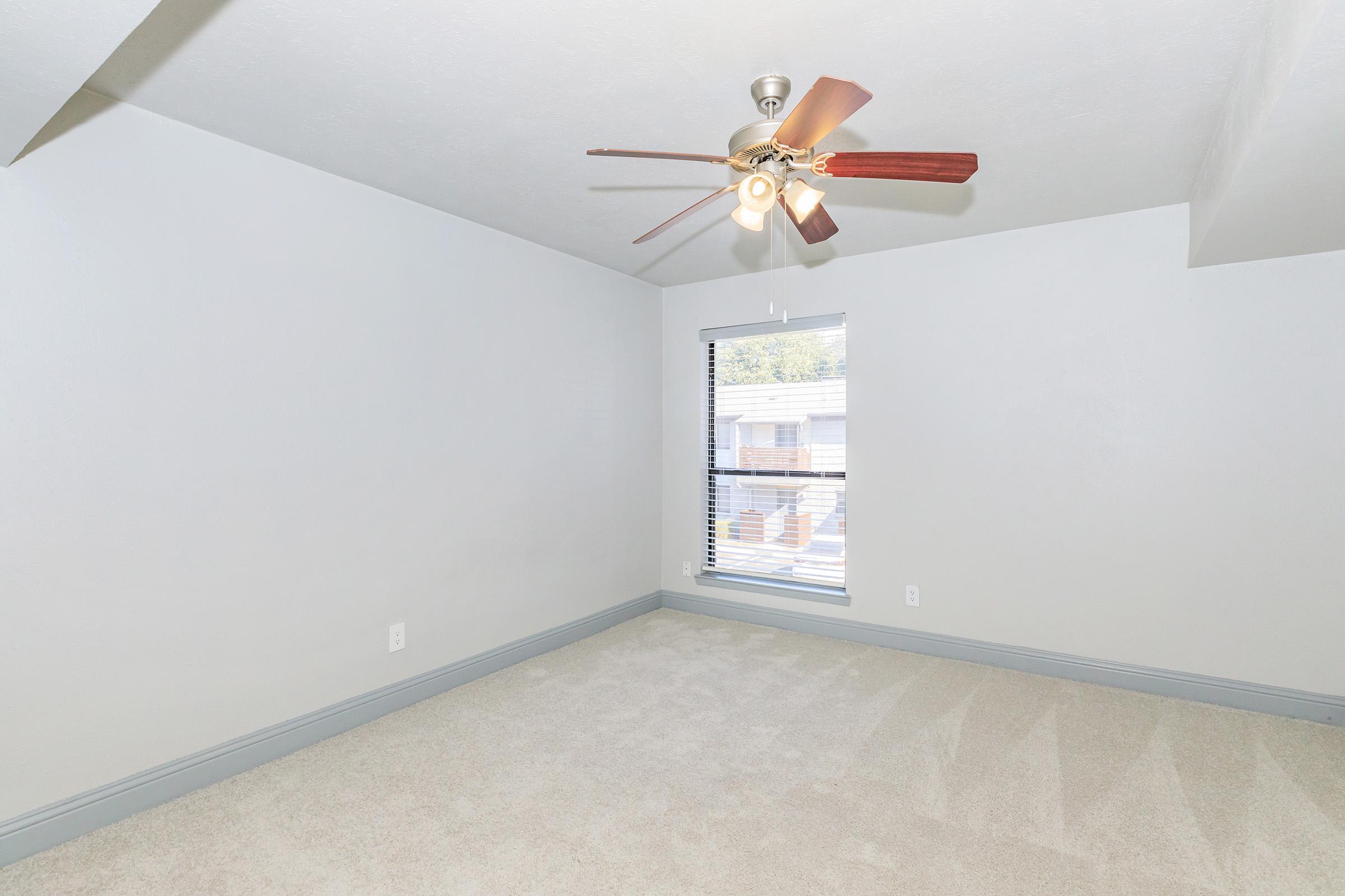 A spacious, empty room with light gray walls and a ceiling fan with wooden blades. The floor is carpeted in a light color, and there is a window with horizontal blinds letting in natural light. The room is well-lit and feels airy, showcasing a simple and clean design.