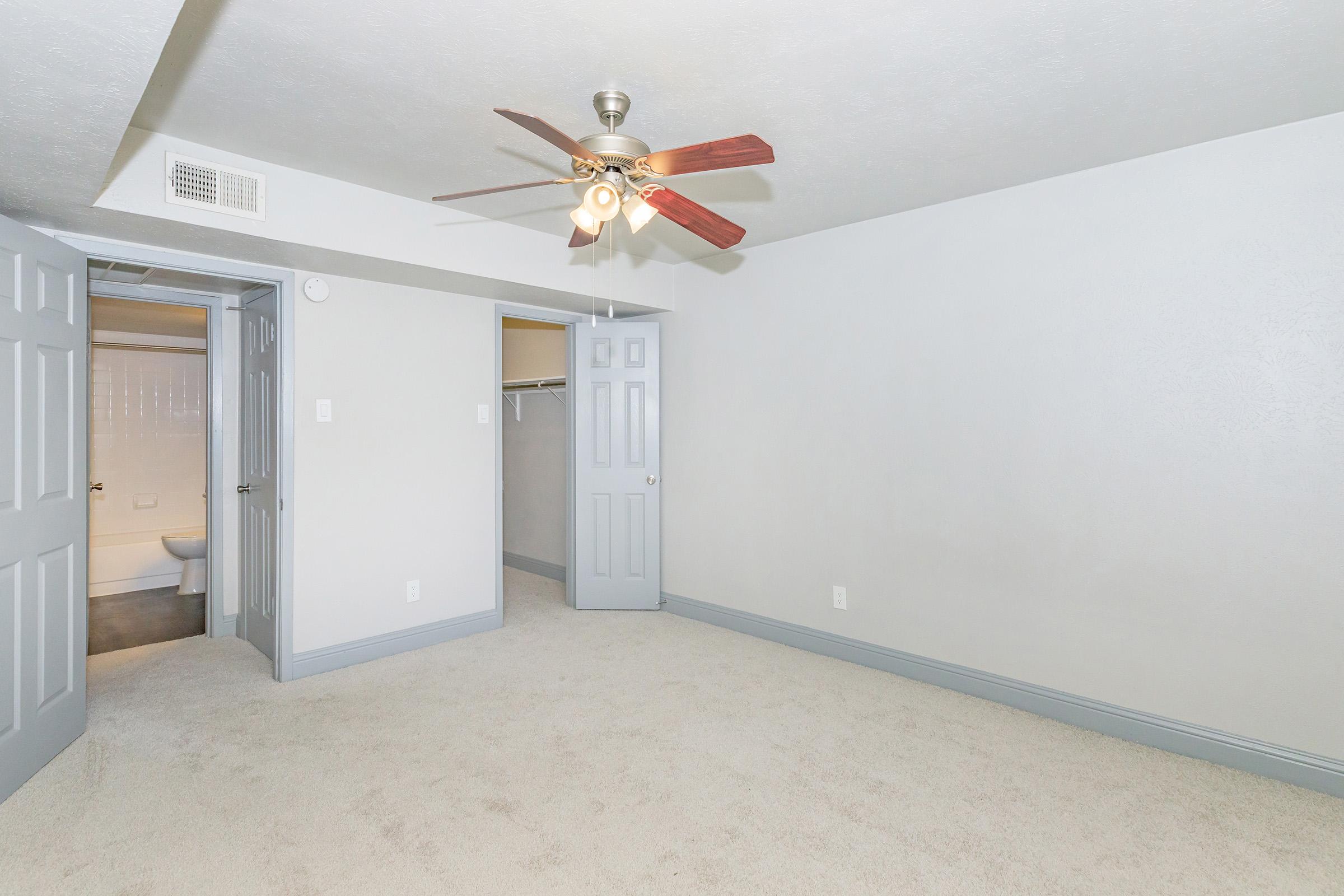 A spacious, light-colored room featuring a ceiling fan with wooden blades. There are two grey doors: one leading to a closet and the other to a bathroom. The floor is carpeted with a soft, neutral tone, and the walls are painted a light shade, creating a welcoming and bright atmosphere.