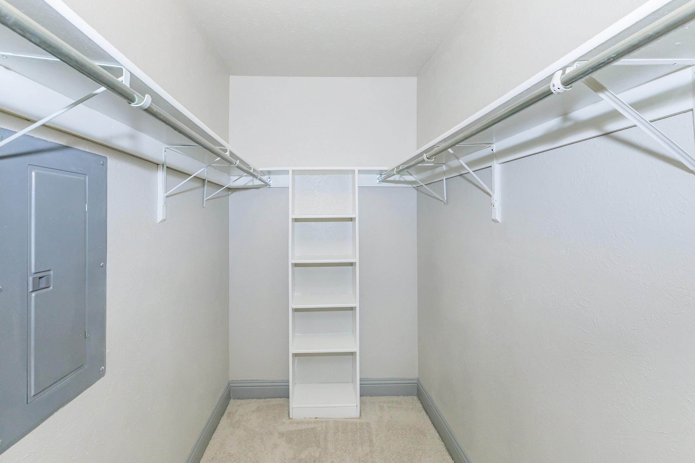 A spacious, minimalist closet with white walls and a light carpet. It features two hanging rods on either side and a central white shelf unit for storage. A gray electrical panel is visible on the left wall, adding functional touches to the uncluttered design.