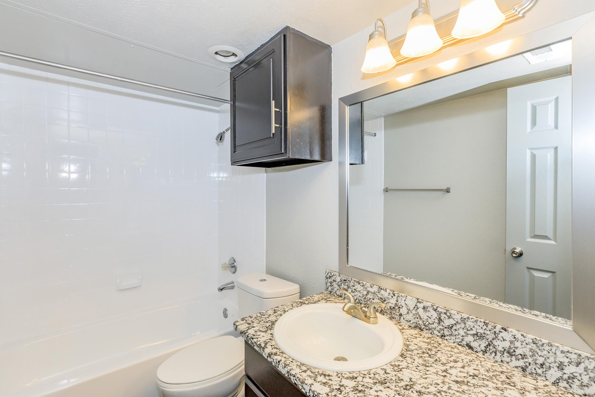 A modern bathroom featuring a white bathtub with a shower, a dark wooden cabinet above the toilet, a granite countertop with a sink, and a large mirror. There are three light fixtures above the mirror, and a closed bathroom door is visible in the background.