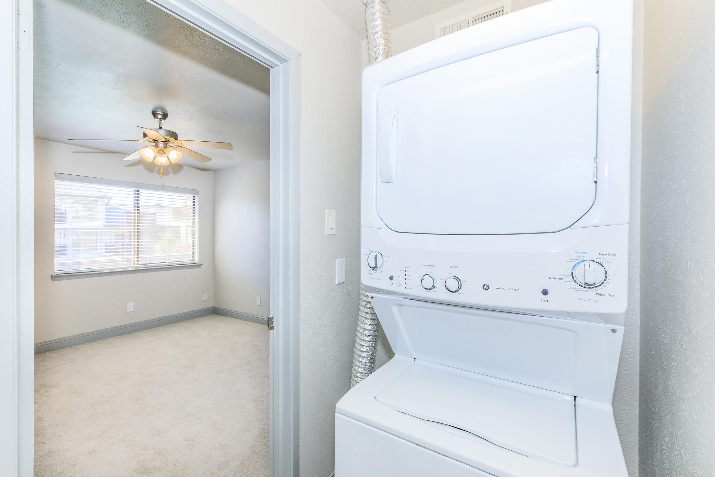 A stackable washer and dryer are positioned in a well-lit laundry area, with a doorway leading to a room featuring a ceiling fan and natural light from windows. The walls are painted light colors, and the carpet is beige.