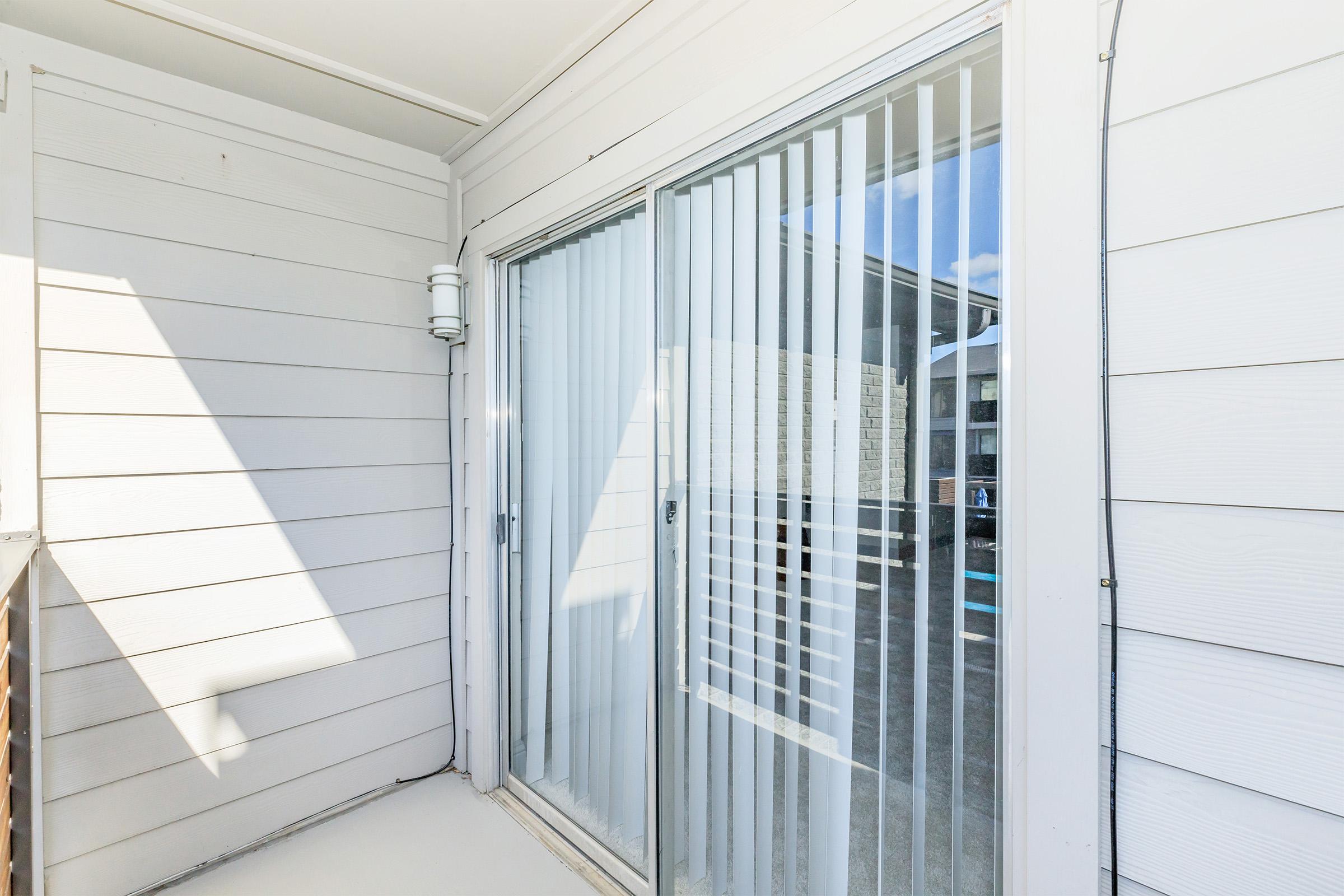 Balcony view featuring sliding glass doors with vertical blinds, light-colored wood siding, and a sunlit outdoor space. The background shows a glimpse of a pool area and a building structure outside.