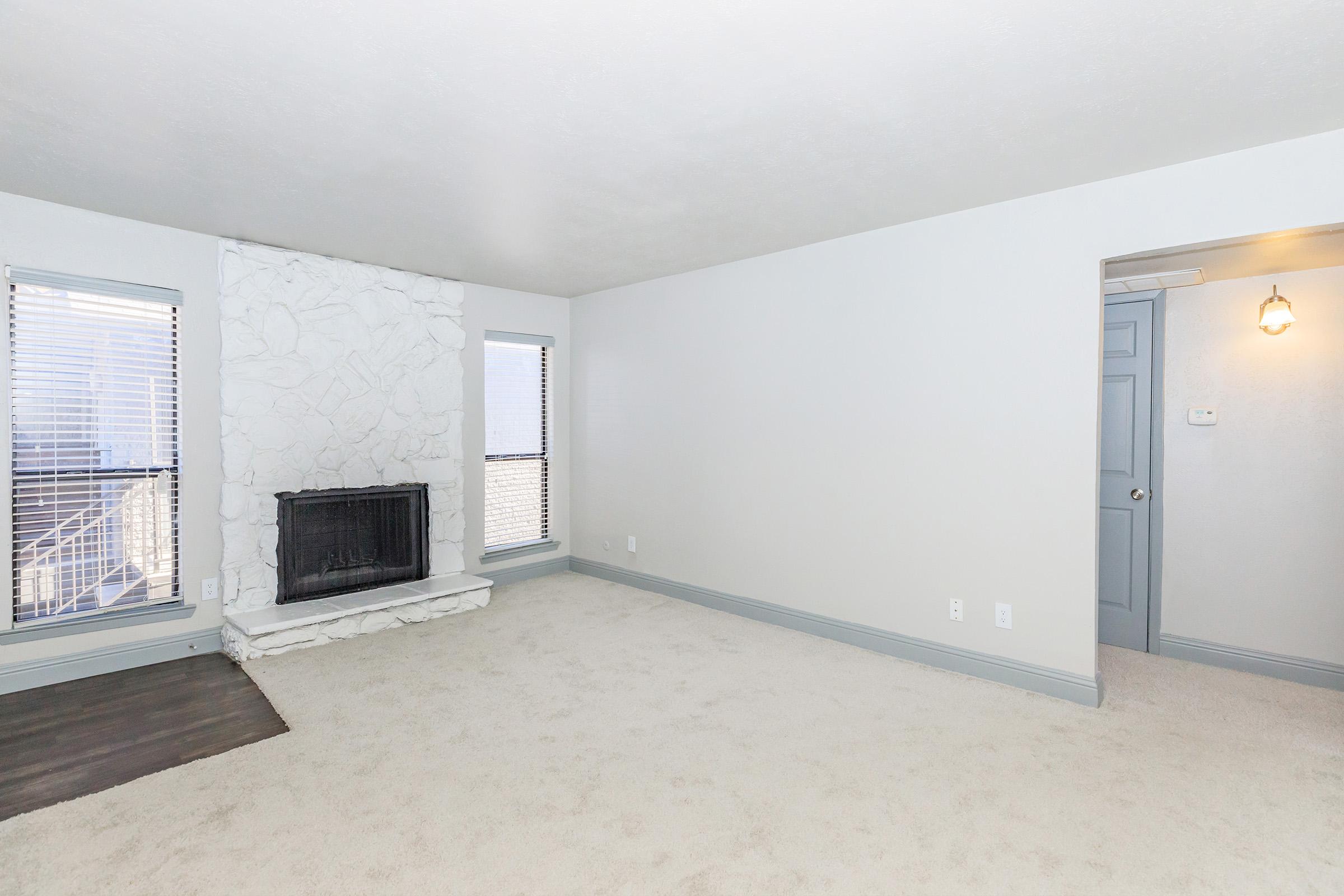 A spacious, well-lit living room featuring a white stone fireplace, beige carpet, and light gray walls. There are two windows with blinds allowing natural light, and a door on the right leading to another area. The room has a clean and modern aesthetic.