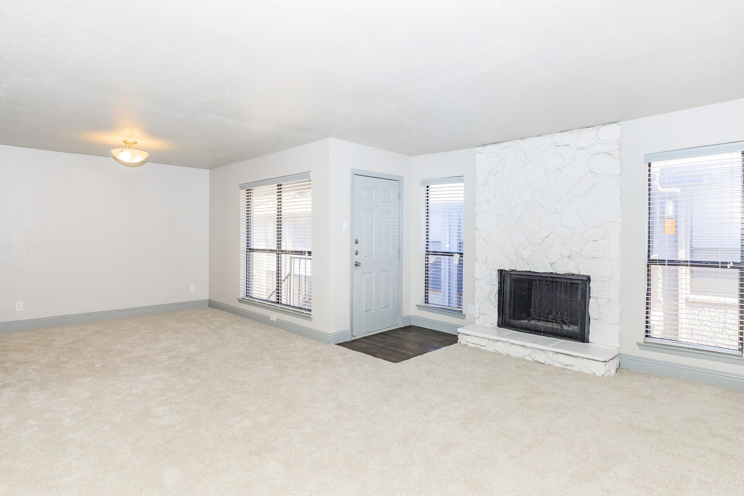 Spacious living room with beige carpet, a white stone fireplace, and large windows allowing natural light. Features a neutral color palette with a simple ceiling light and entry door.