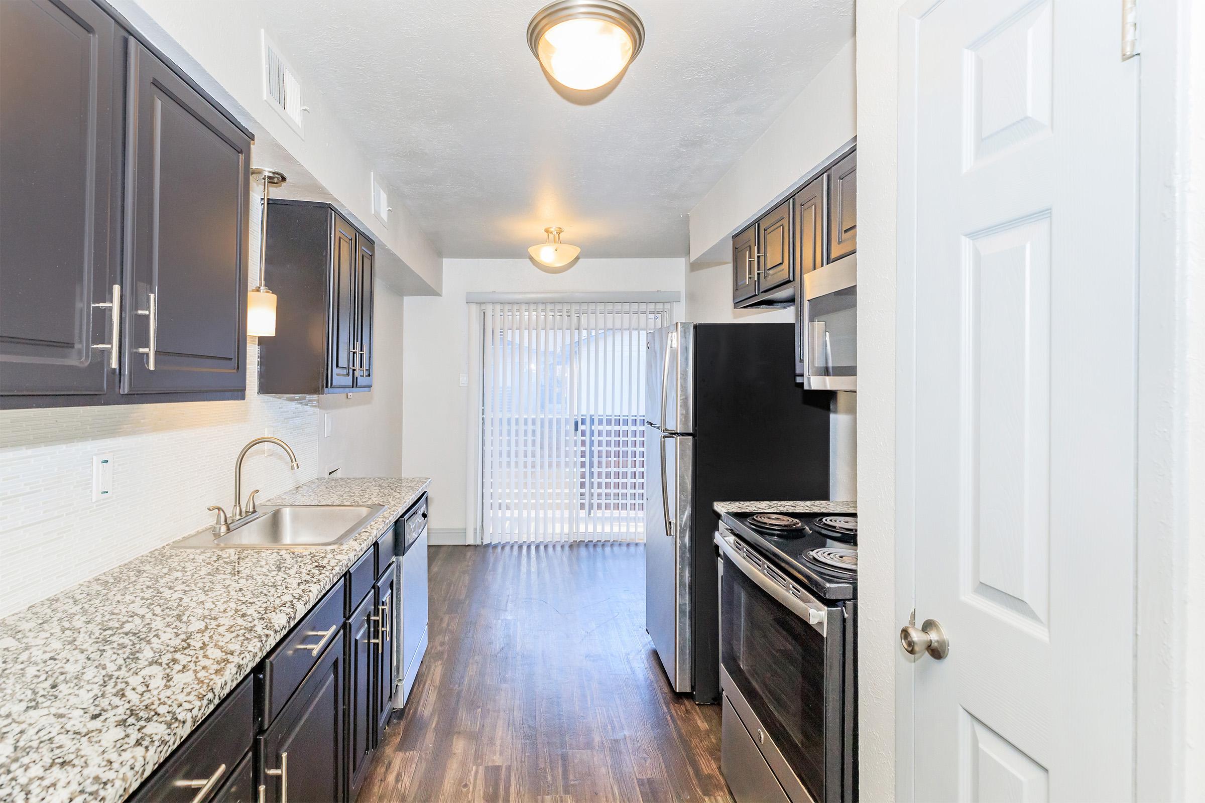 A modern kitchen featuring dark cabinets, a granite countertop, and stainless steel appliances. The space includes a sink, stove, and a refrigerator. Natural light enters from a sliding glass door leading to a balcony with white blinds. The floor is dark wood, creating a contemporary aesthetic.