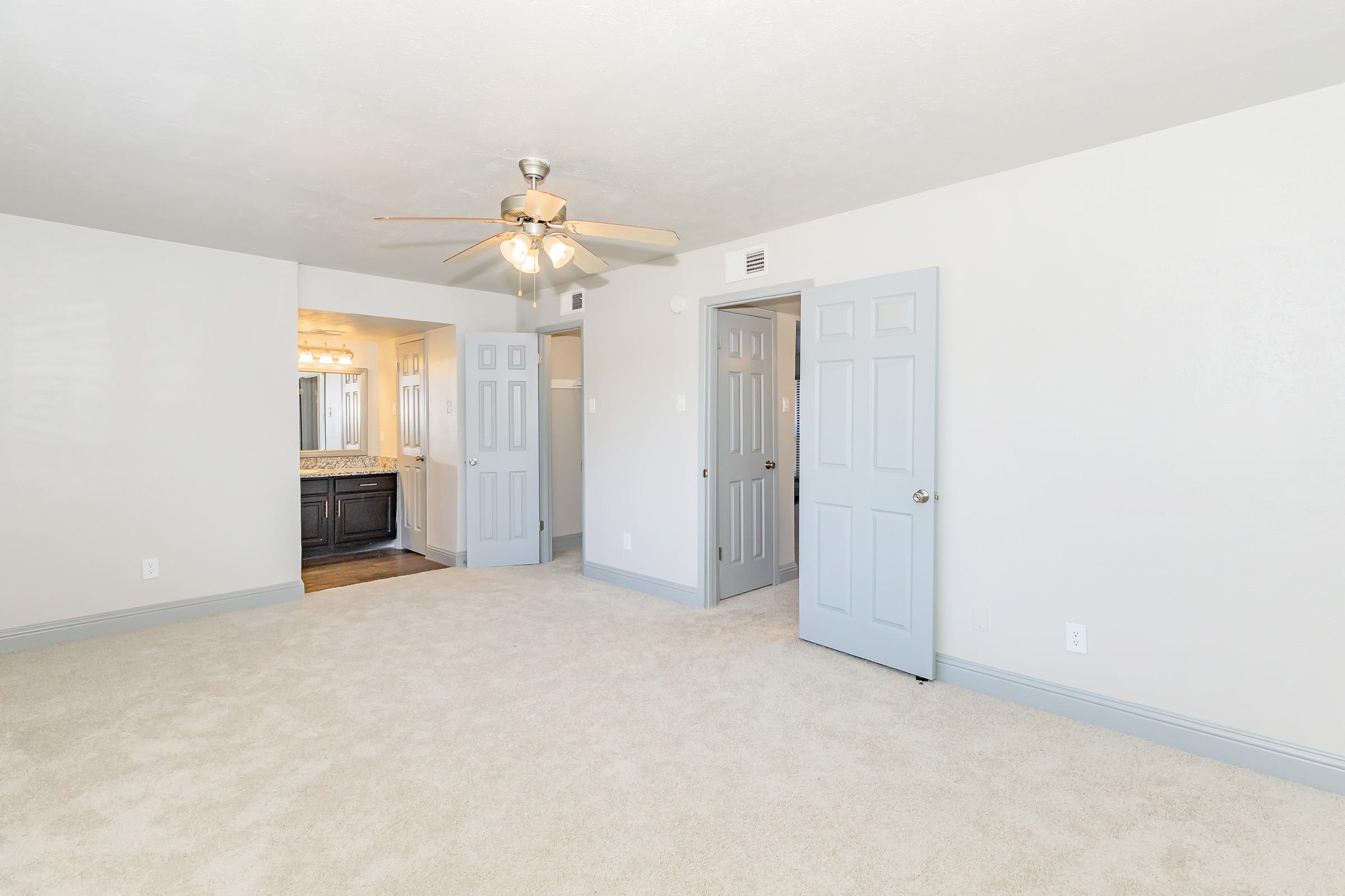 A spacious room with light-colored walls and plush carpet. A ceiling fan is installed above, and there are two closed doors leading to other areas. One door on the left opens to a bathroom with a dark vanity. Natural light illuminates the space, creating a bright and inviting atmosphere.
