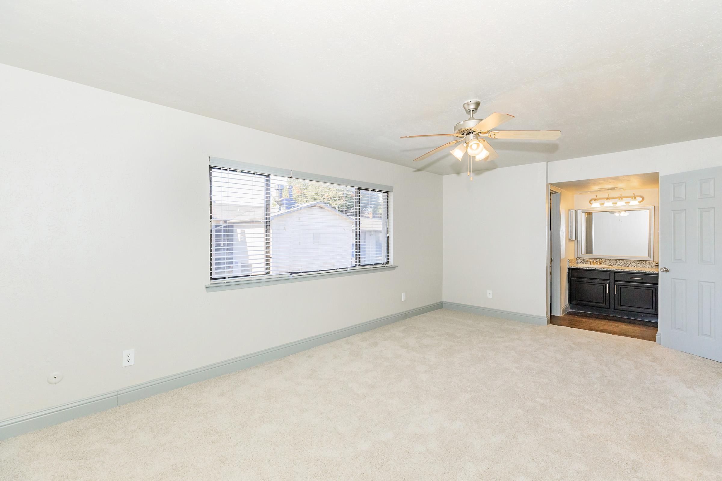 A well-lit, empty bedroom featuring light-colored walls and carpet. There is a ceiling fan with wooden blades and a large window with blinds, providing natural light. To the right, there's a doorway leading to a bathroom with a double sink and dark cabinetry. The space feels open and inviting.