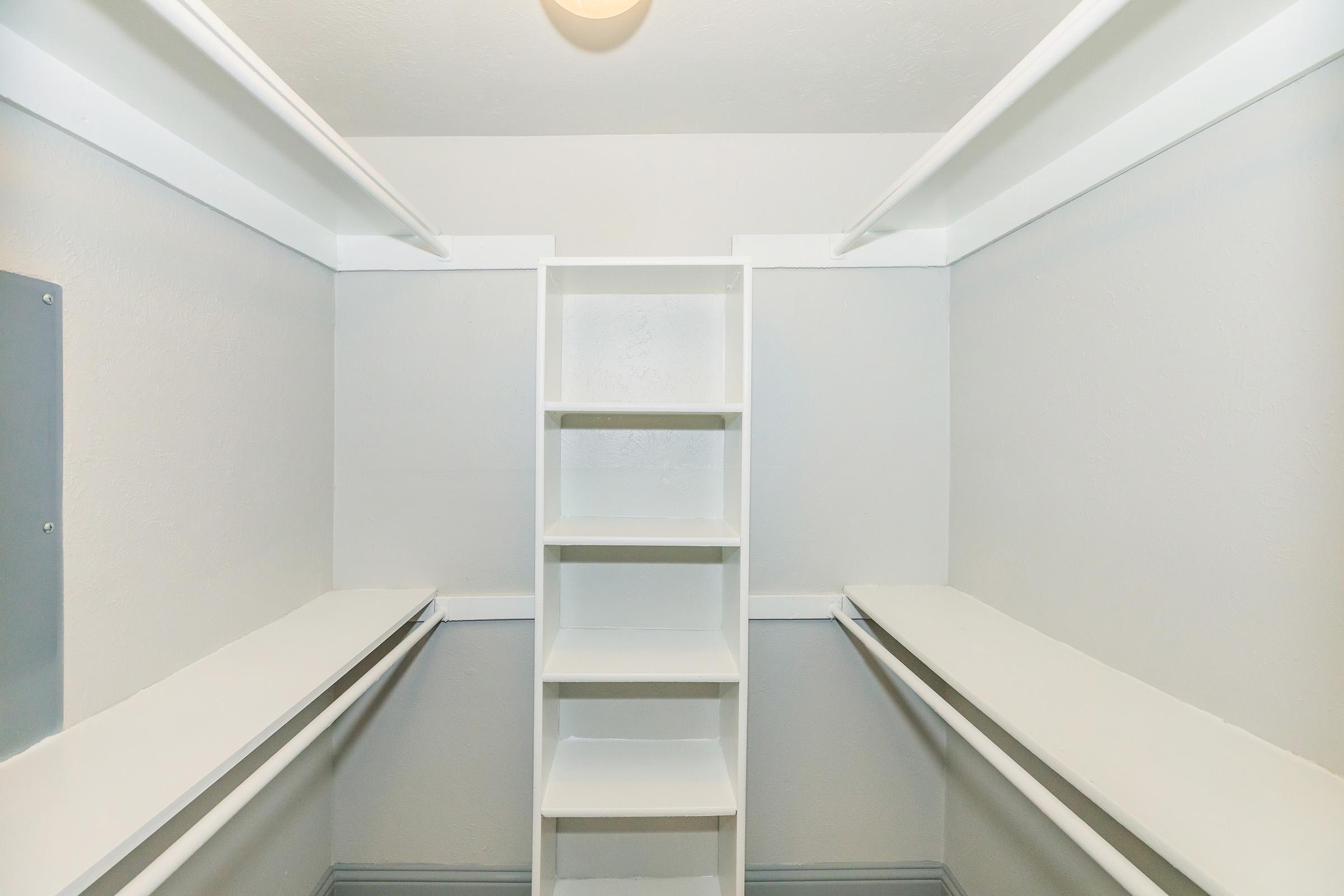 A clean, empty closet with white walls and shelving. The closet features a tall, narrow shelf in the center and two horizontal shelves on each side, providing storage space. The lighting is soft and bright, creating a spacious feel. Ideal for organization and storage solutions.
