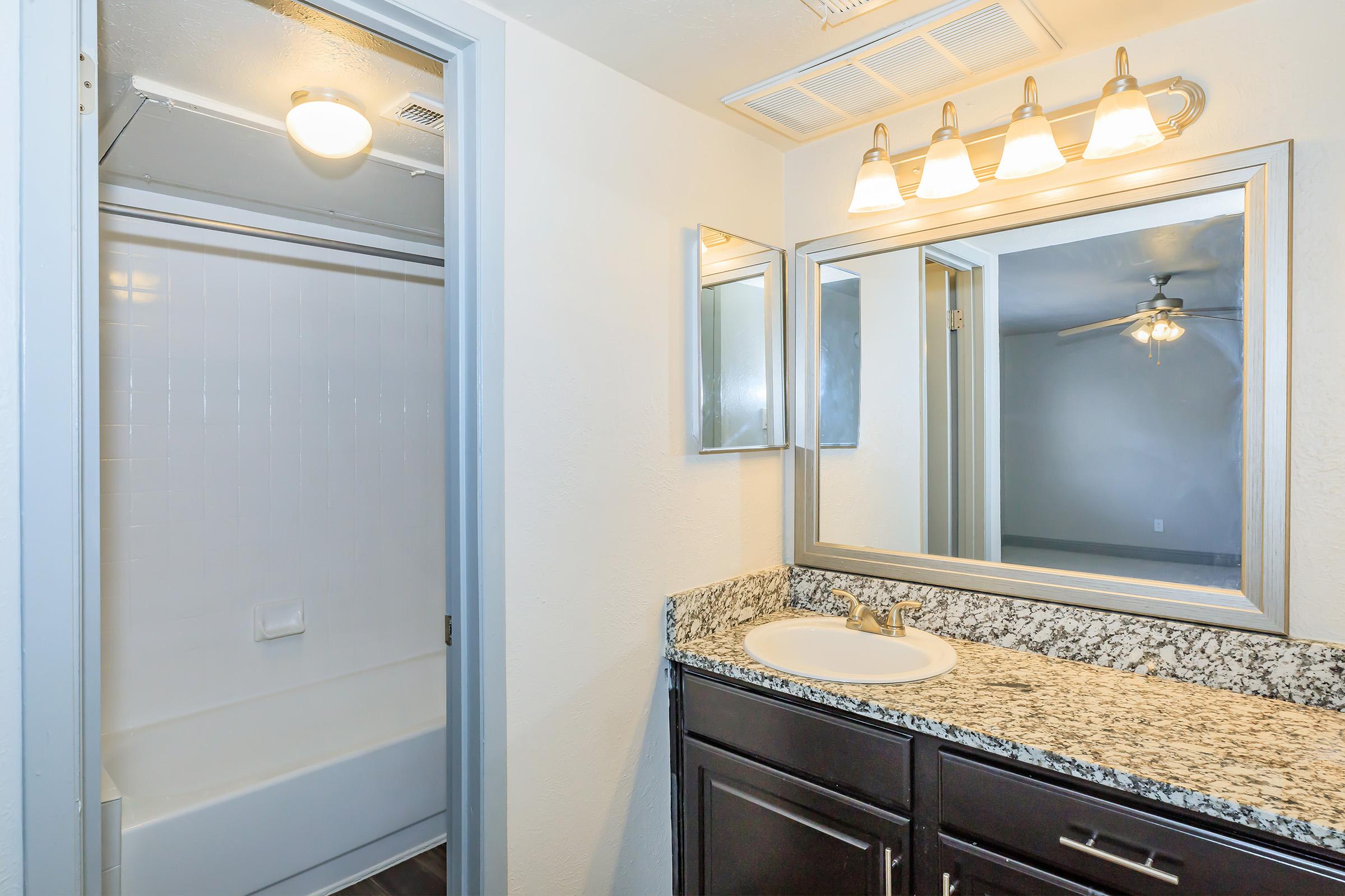 A modern bathroom featuring a granite countertop with a sink, a large mirror, and a light fixture above. To the left, there's a shower/tub combo with a white curtain rod. The walls are painted in neutral tones, and the space is well-lit with natural and artificial light.