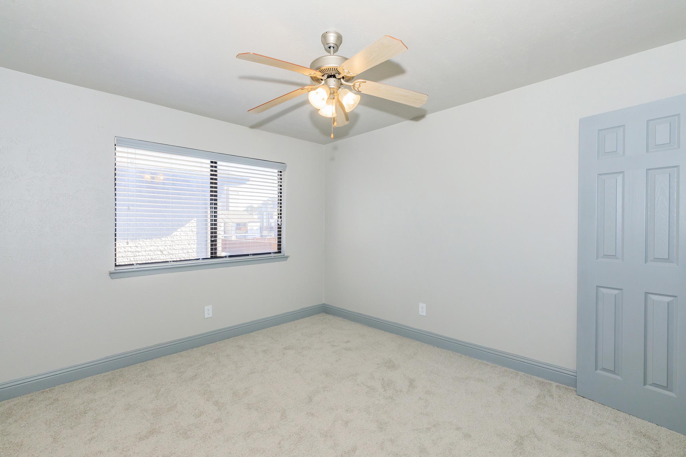 A well-lit room featuring a ceiling fan with light fixtures, a window with blinds, and neutral-colored walls. The carpeted floor is light beige, and there is a gray door on the right side. The overall atmosphere is clean and inviting, suitable for various purposes such as a bedroom or office.
