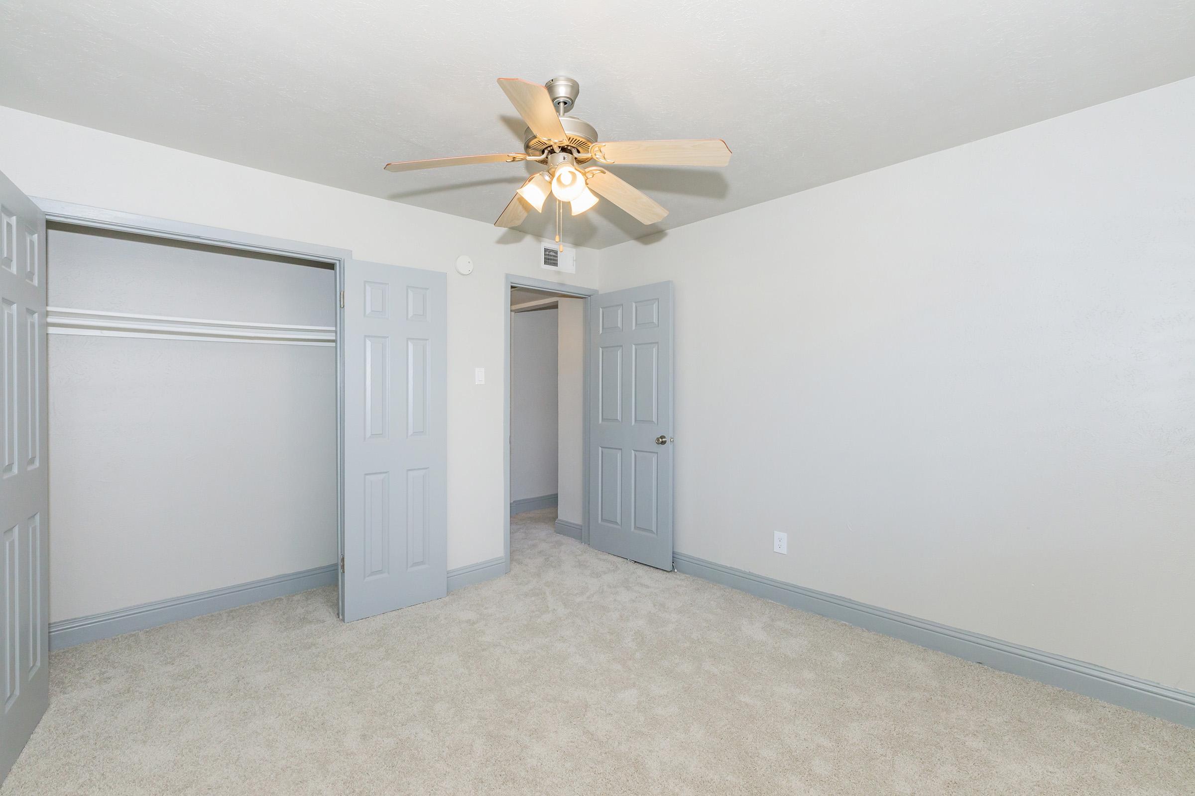 A clean, neutral-toned bedroom featuring a ceiling fan with light, light gray walls, and carpeted flooring. Two gray doors lead to a closet and another room. The space has an airy feel with good lighting and minimal decor, creating a blank canvas for personalization.