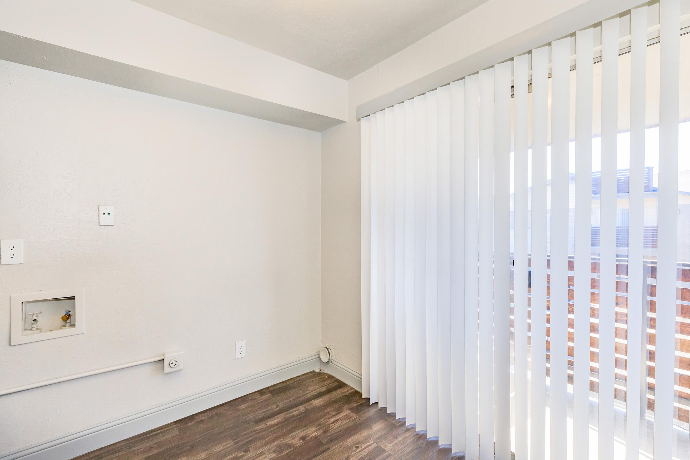 A bright, empty room with light gray walls and wooden flooring. There are vertical white blinds covering a large window, allowing natural light to enter. An electrical outlet is visible on the wall, and a small recessed area is present near the floor. The overall ambiance is minimalistic and modern.