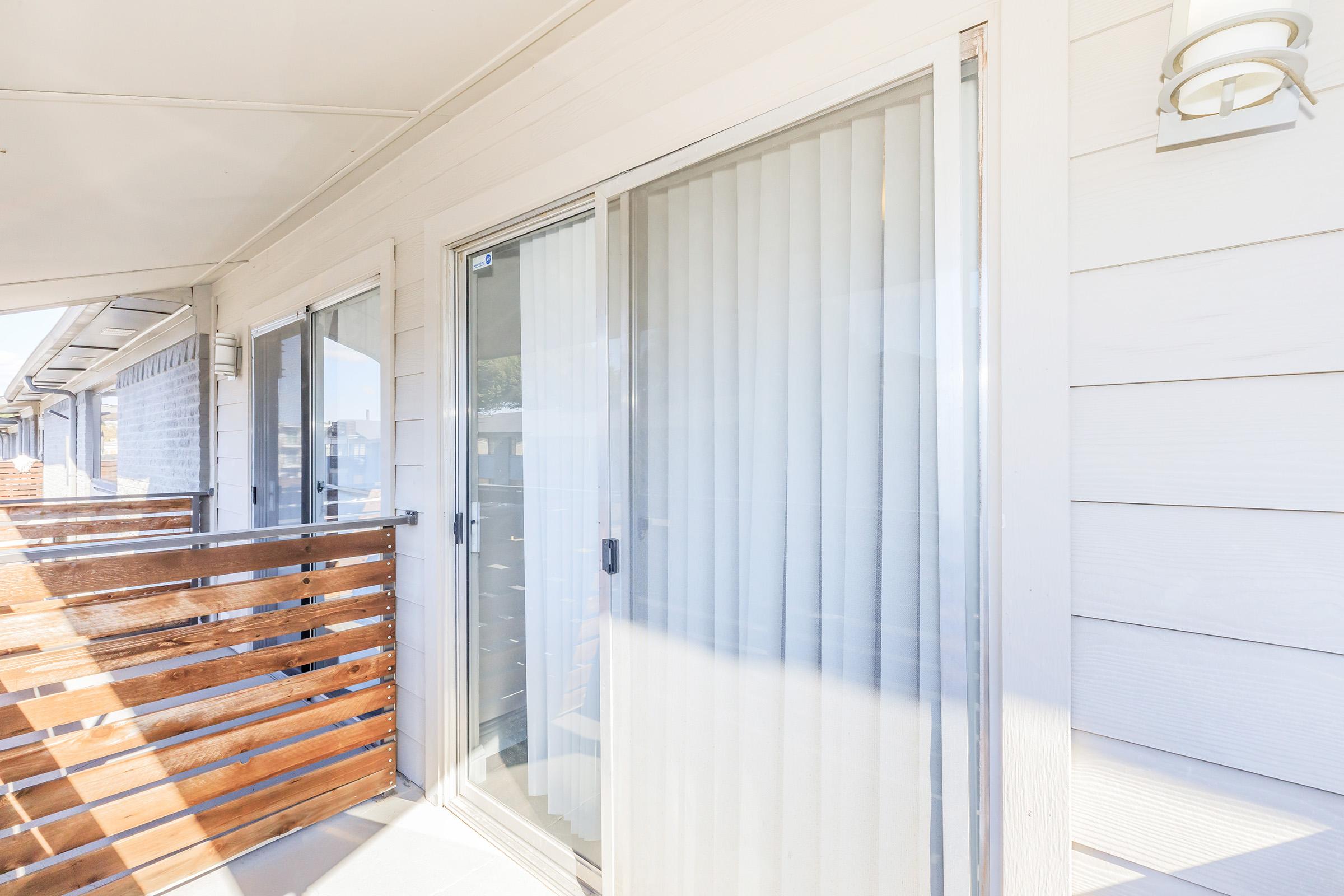 A patio door with vertical blinds, set within a light-colored exterior wall. A wooden railing is visible in the foreground, with the patio area appearing bright and inviting. The scene captures a modern and clean aesthetic typical of a residential outdoor space.
