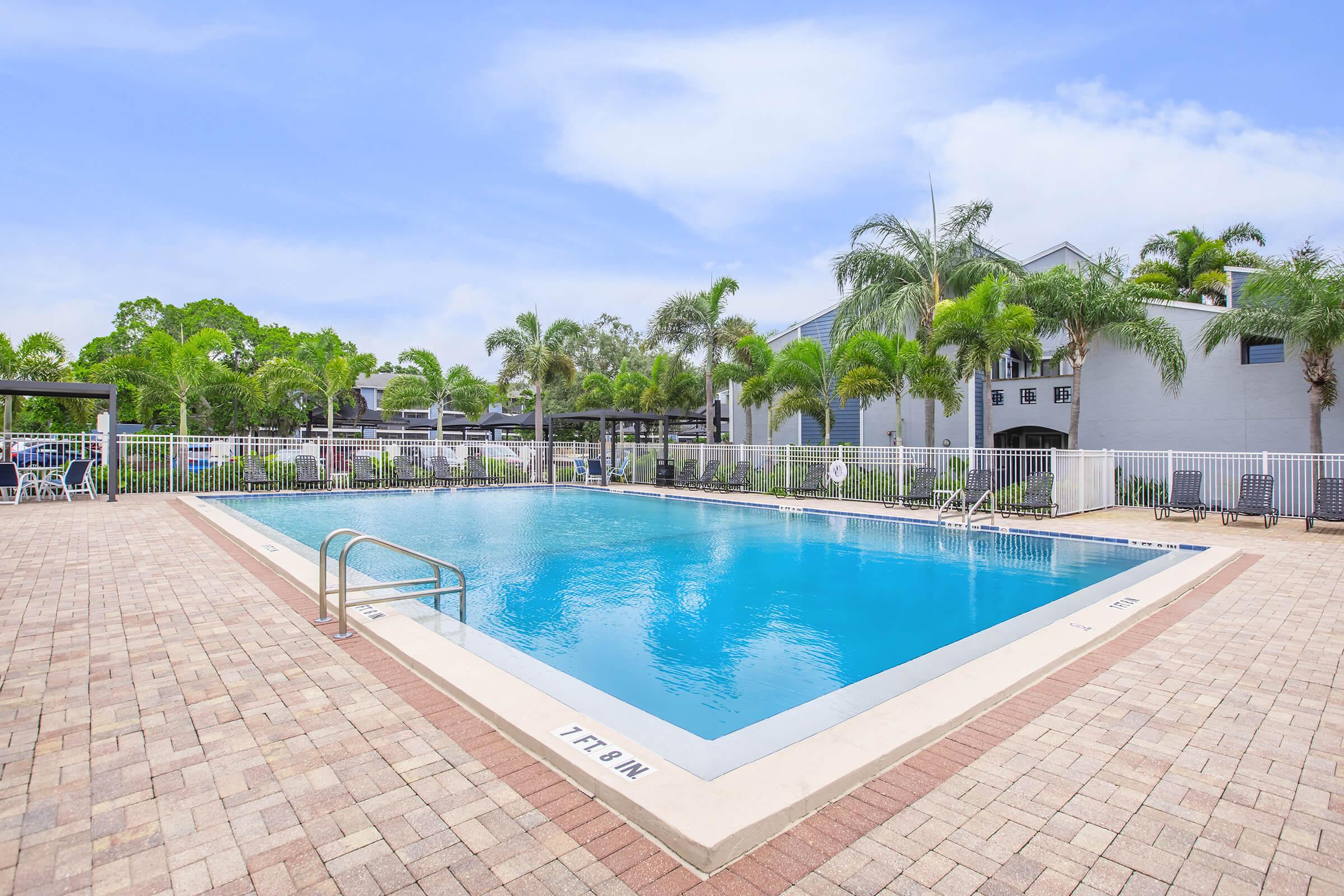 A clear blue swimming pool surrounded by palm trees and lounge chairs, set in a well-maintained outdoor area. The pool's shallow end is marked, and buildings are visible in the background, creating a relaxing atmosphere. The sky is bright with a few clouds.
