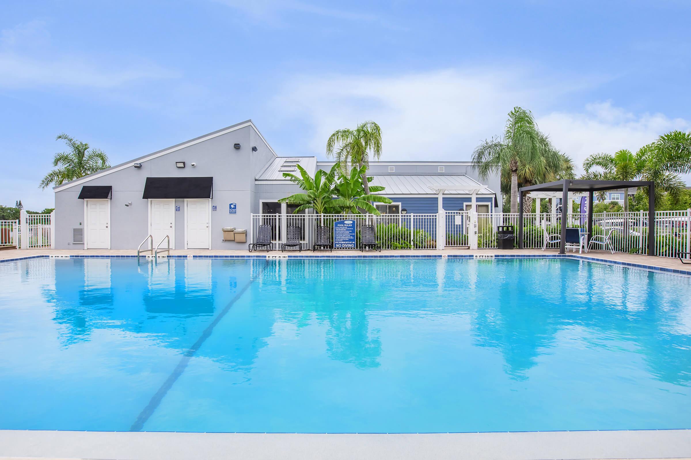 Swimming pool with clear blue water, surrounded by palm trees and lush greenery. In the background, there is a building with a light gray exterior and outdoor seating areas. The scene is under a bright blue sky, creating a serene and inviting atmosphere.