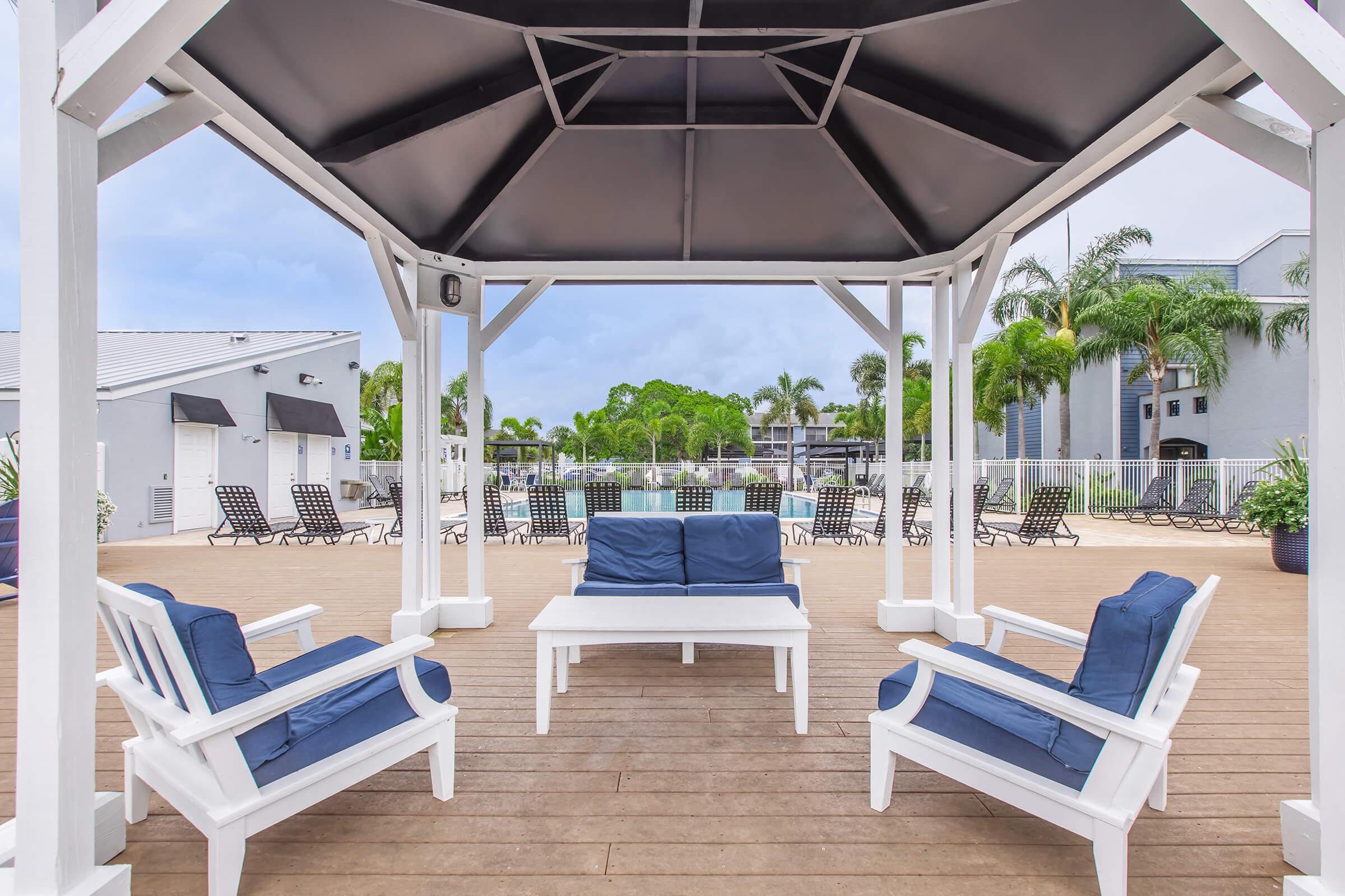A shaded outdoor seating area with white furniture, including a couch and chairs with blue cushions, under a gazebo. In the background, there is a pool area surrounded by palm trees and lounge chairs, set against a clear sky. Perfect for relaxation in a sunny environment.