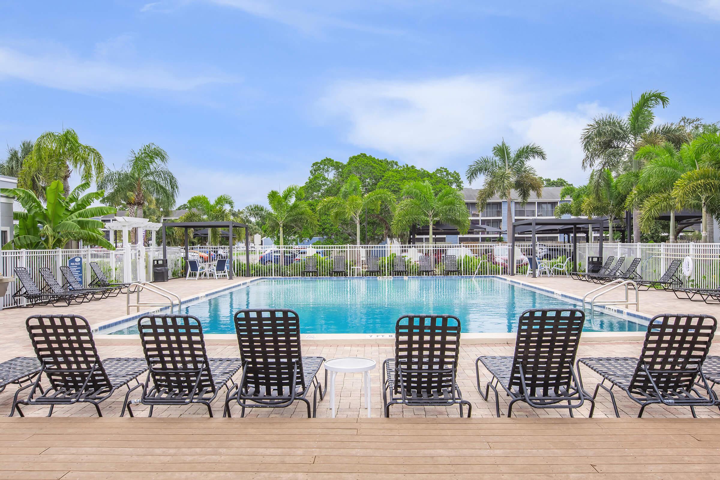 A pool area with a clear blue swimming pool surrounded by lounge chairs. Lush greenery and palm trees are visible in the background, creating a tropical atmosphere. A few buildings and umbrellas are present, typical of a resort or apartment complex setting. The sky is bright and sunny.
