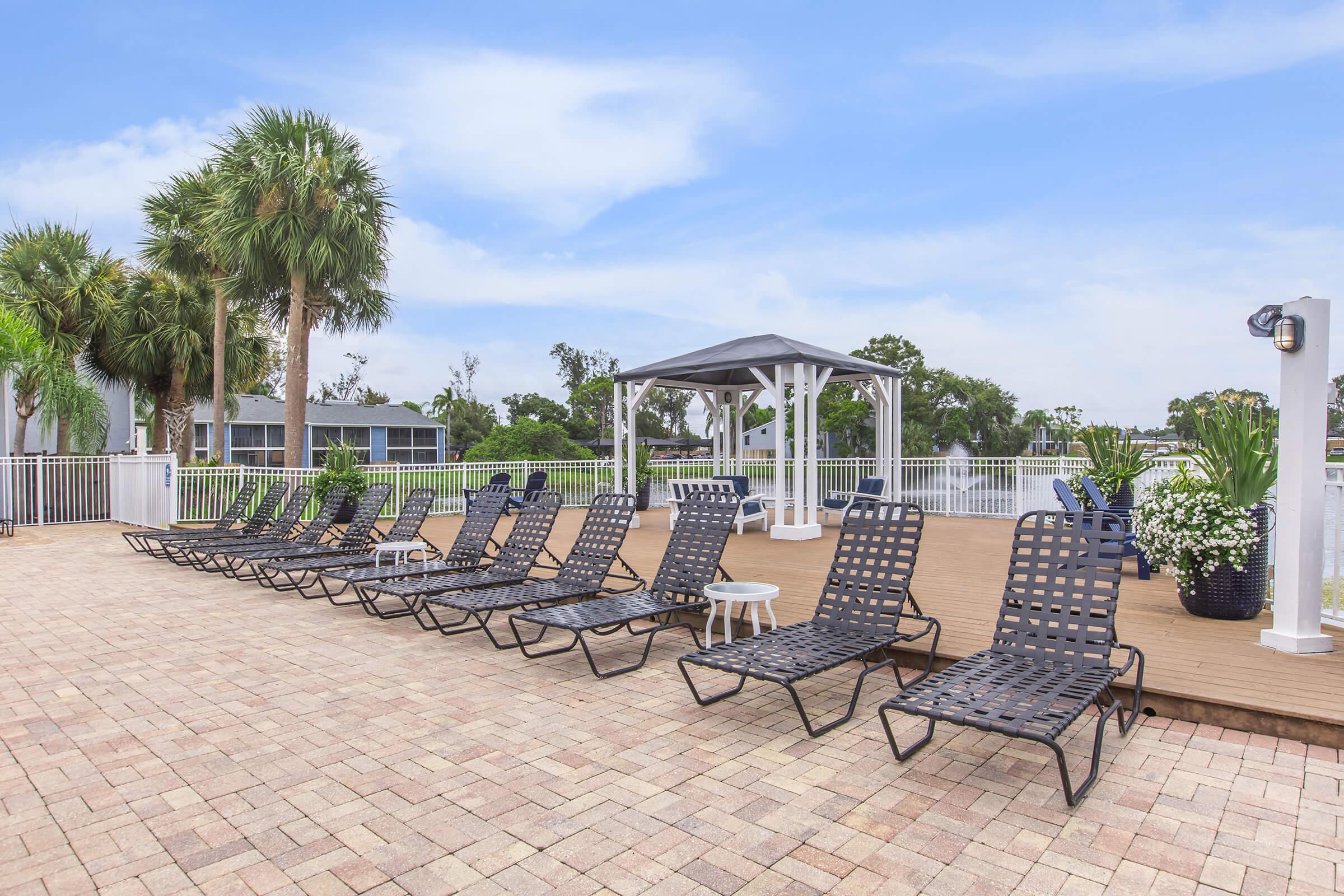 A sunny outdoor patio area featuring multiple black lounge chairs arranged in a row. In the background, there are palm trees and a gazebo with white railings. The ground is paved with light-colored bricks, and potted plants add greenery to the scene. A blue sky with scattered clouds completes the relaxing atmosphere.