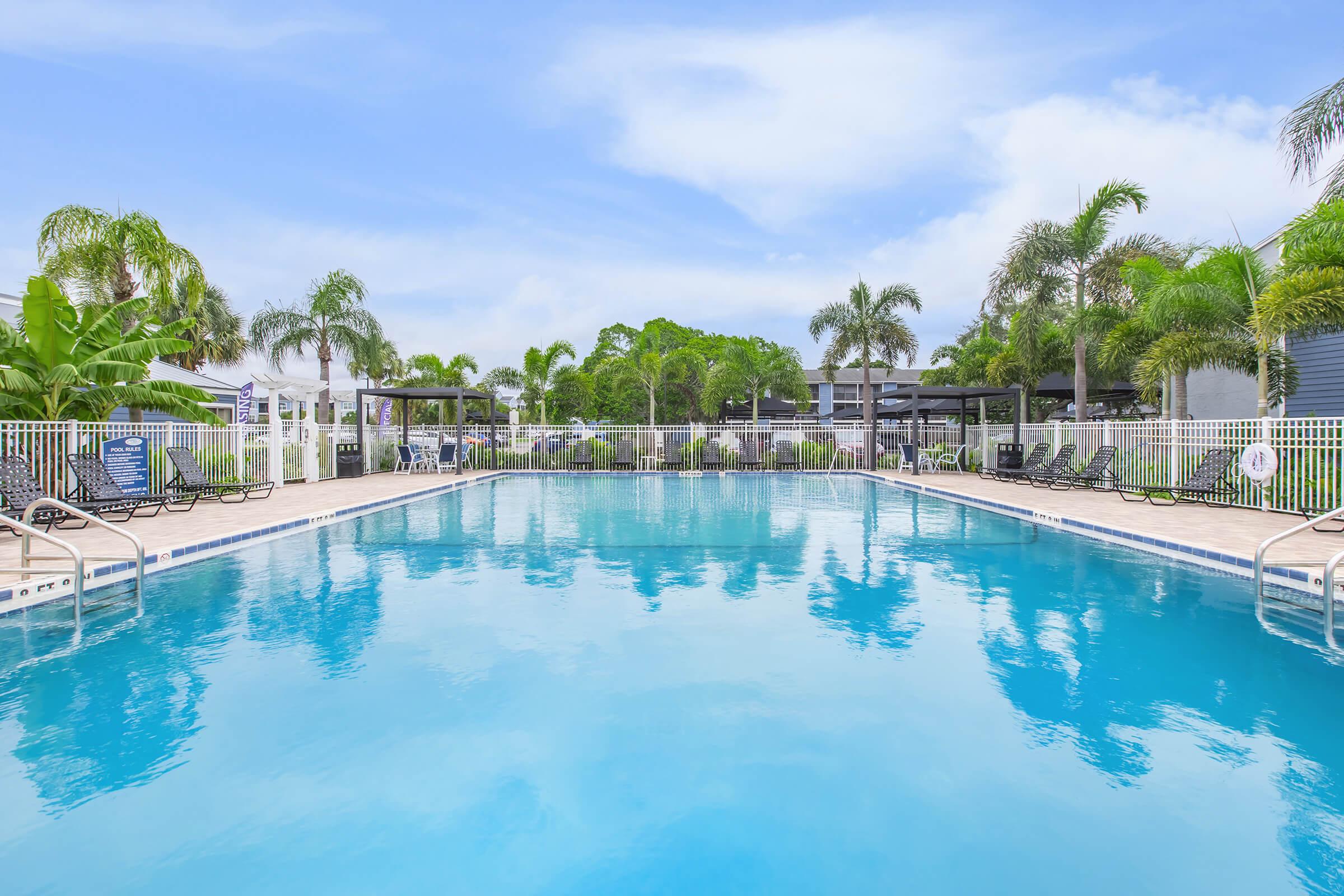 A clear blue swimming pool surrounded by palm trees and lounge chairs, with a light blue sky in the background. The pool area features white fencing and shaded cabanas, creating a relaxing atmosphere for recreation and leisure.