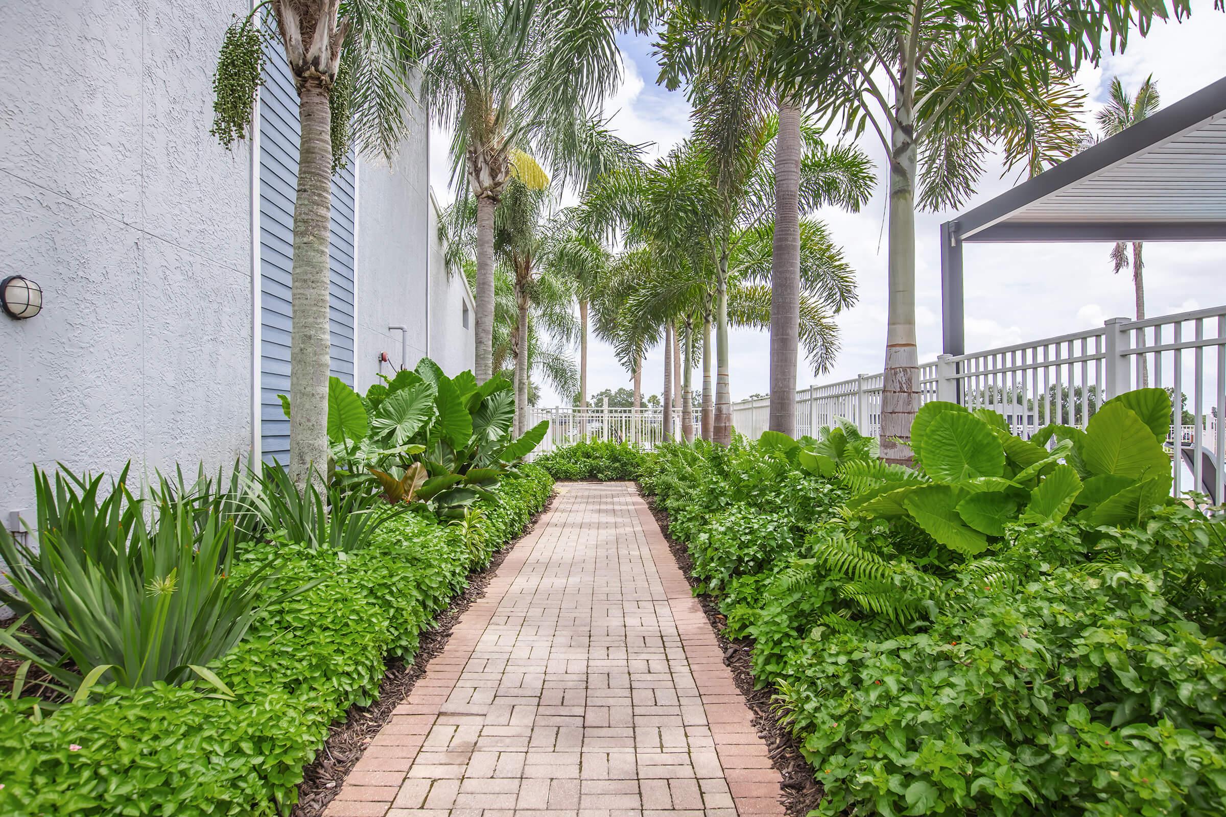 A well-maintained pathway lined with vibrant green plants and palm trees, leading through a landscaped garden. The path consists of beige paving stones bordered by lush foliage on both sides, creating a serene and inviting atmosphere, with a cloudy sky in the background.