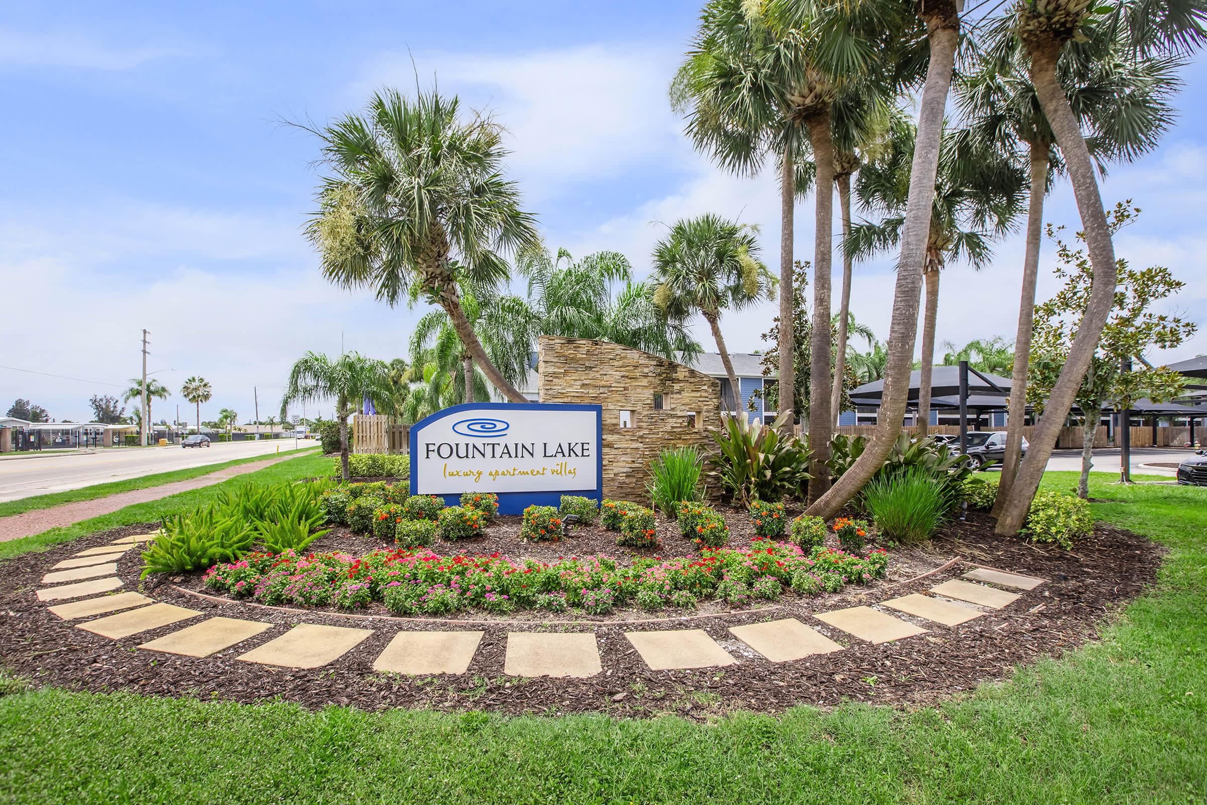 A well-manicured landscape featuring a sign for Fountain Lake luxury apartment homes, surrounded by vibrant flowers, palm trees, and lush greenery. The area is neatly maintained with stone and concrete pathways, set against a clear sky and adjacent to a road.