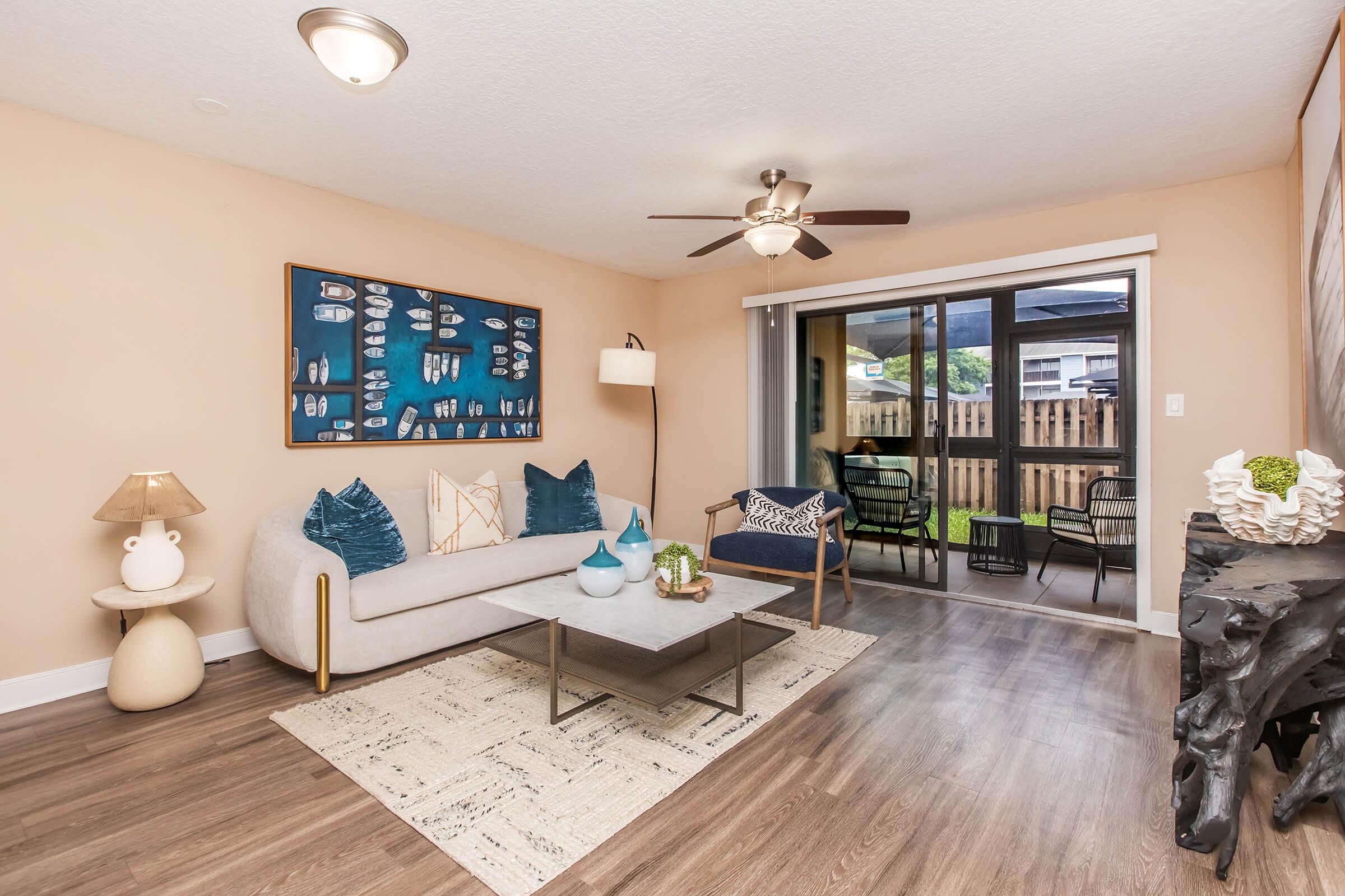 A stylish living room featuring a light-colored sofa with decorative pillows, a coffee table with a plant, and a modern lamp. The space has a blue and white artwork on the wall, hardwood flooring, and a sliding glass door leading to a balcony with outdoor seating. Natural light brightens the room.