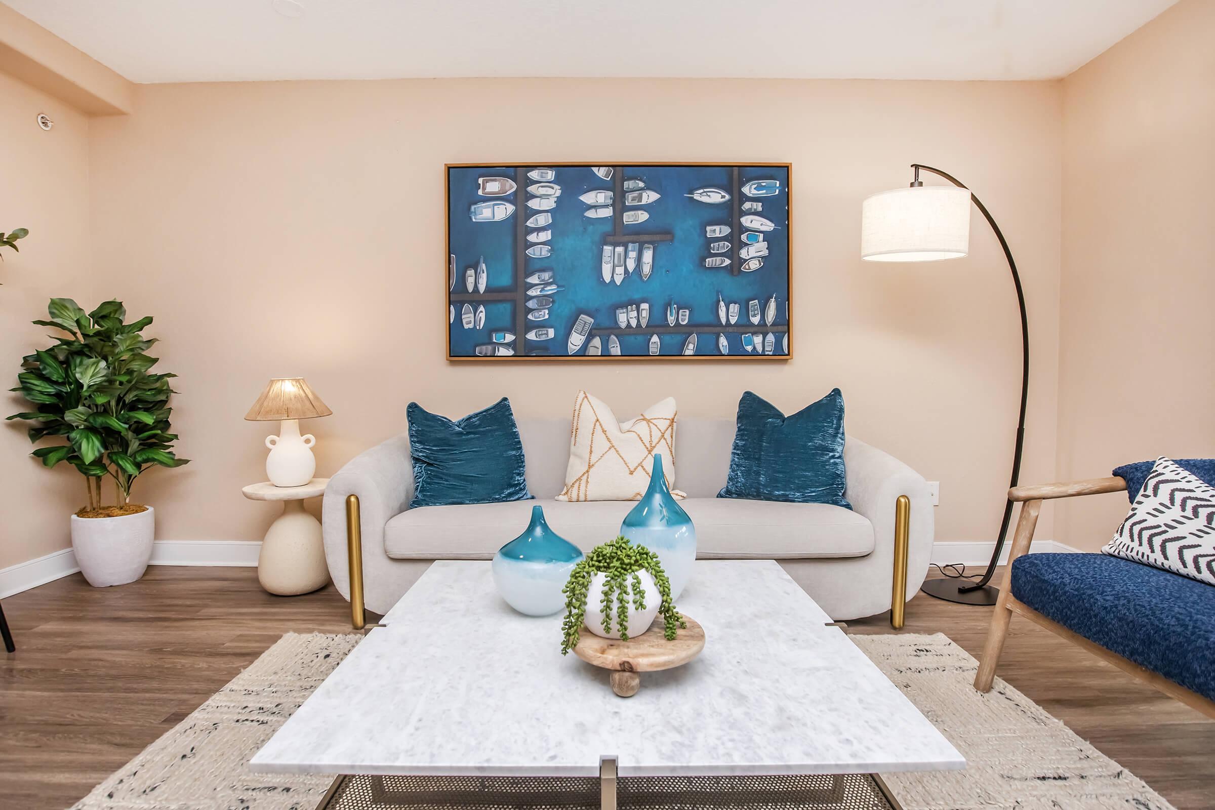 A cozy living room featuring a light-colored sofa with blue and white decorative pillows, a marble coffee table with plants, and a stylish floor lamp. The wall art depicts various boats on a blue background, complemented by a potted plant and soft lighting for a warm atmosphere.