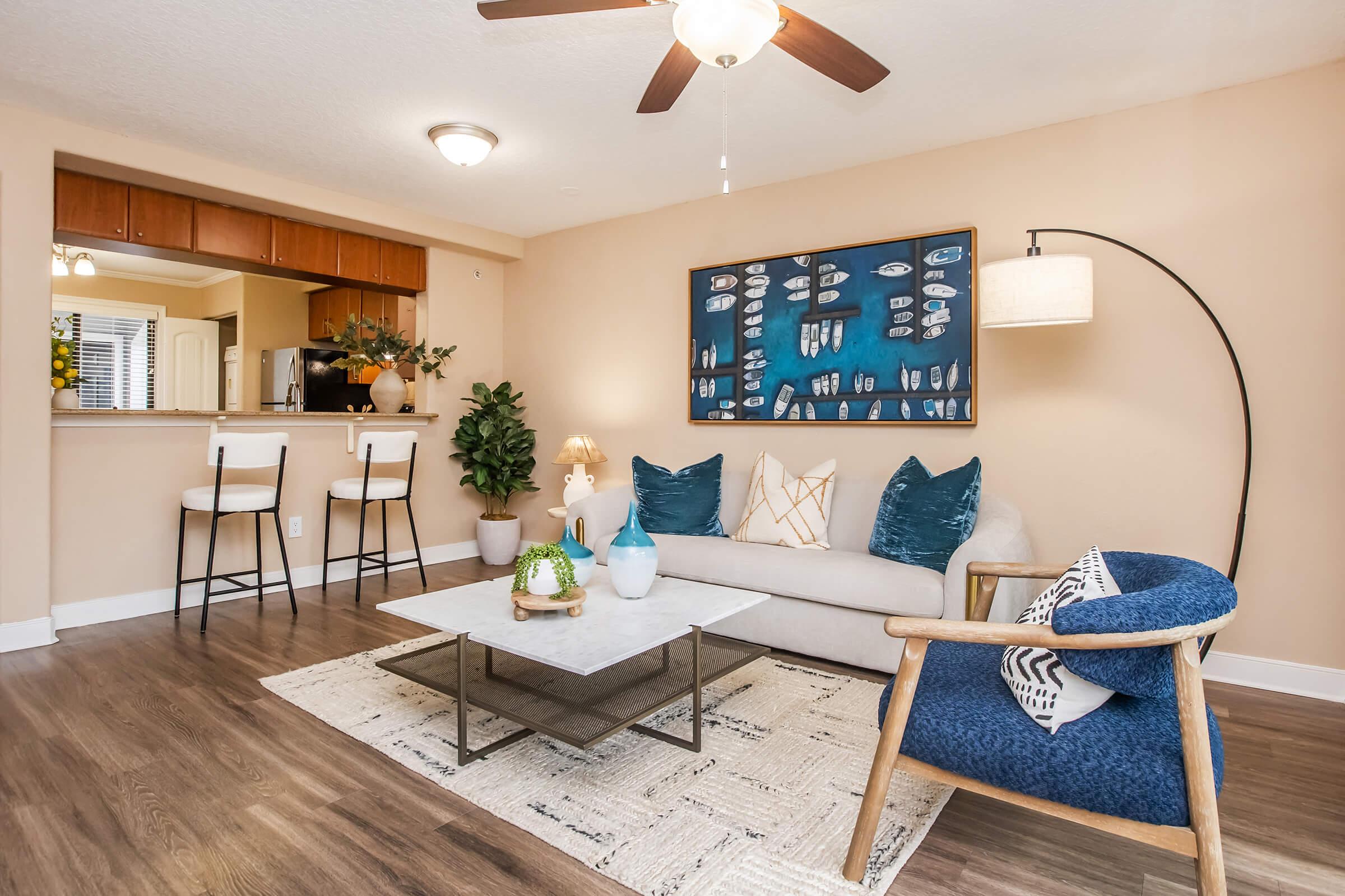 A cozy living room featuring a light-colored sofa with blue and white decorative pillows, a stylish coffee table on a textured rug, and a floor lamp. In the background, there's a small dining area with two bar stools and a kitchen counter. Soft lighting and decorative plants add warmth to the space.