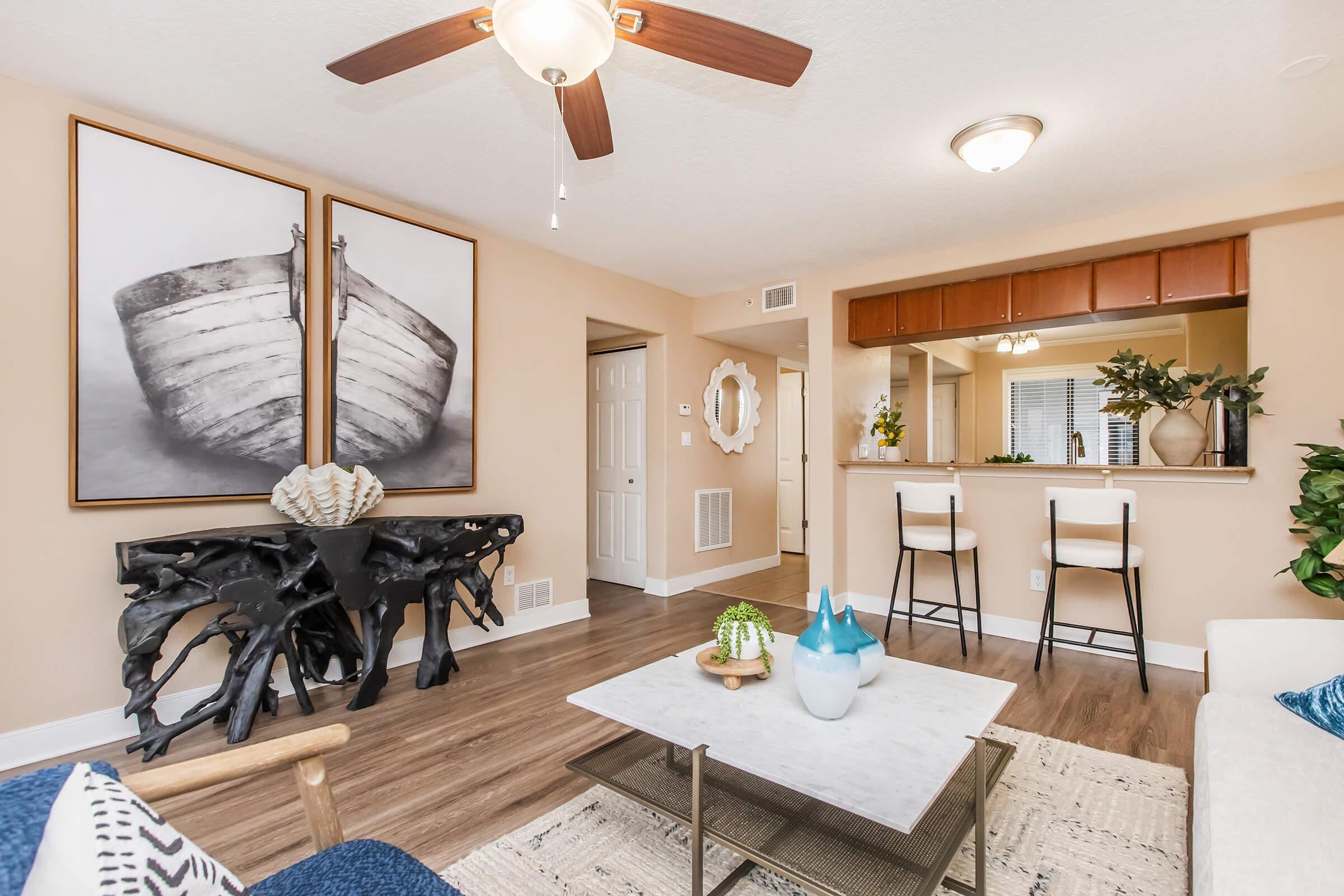 A modern living room featuring a light color palette with a ceiling fan. Two large black and white boat prints hang on the wall, complemented by a decorative console table. A white marble coffee table sits in the center, surrounded by a cozy couch and bar stools at a kitchen counter in the background. Green plants add a touch of nature.