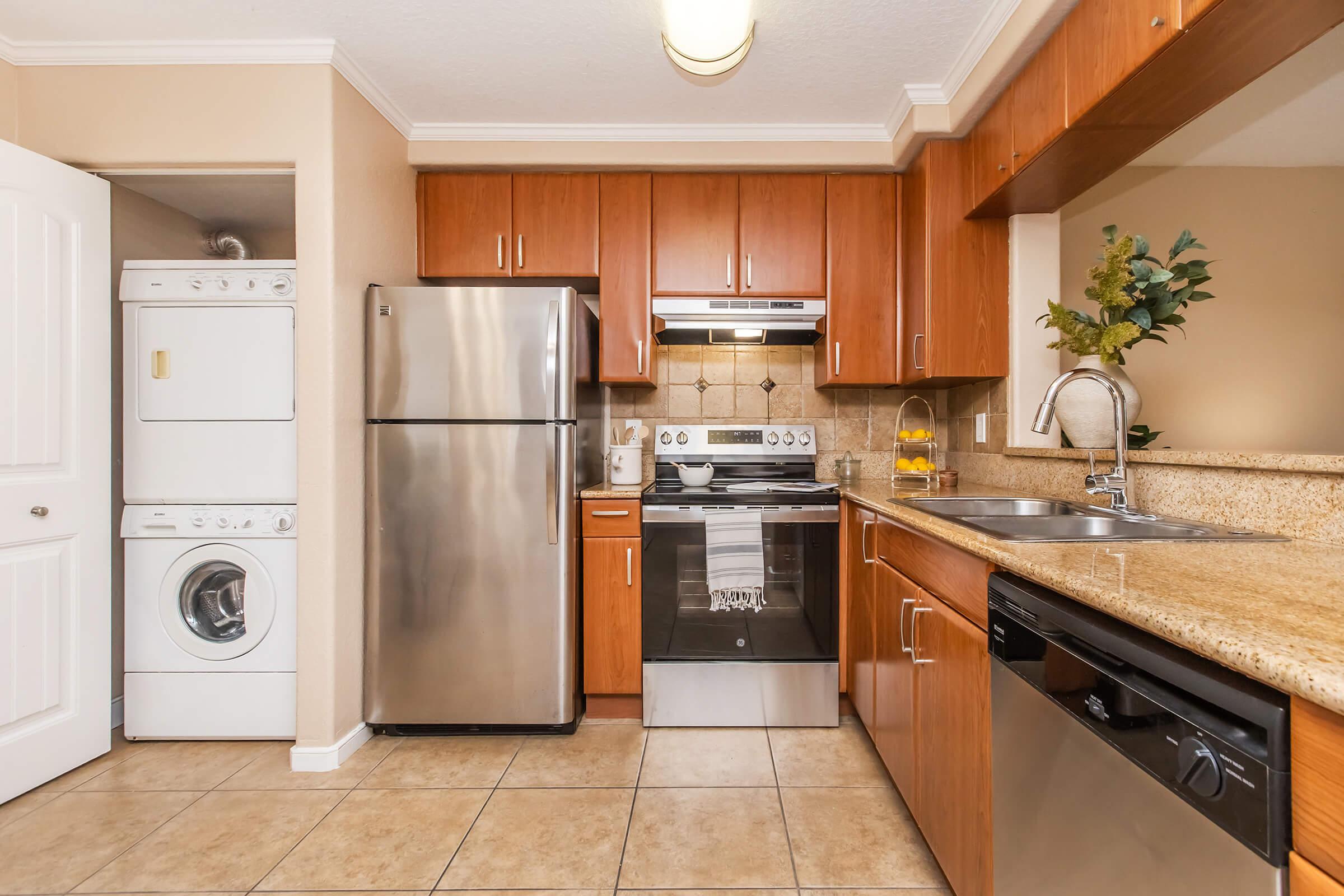 A modern kitchen featuring wooden cabinetry, stainless steel appliances including a refrigerator, stove, and dishwasher, granite countertops, and a washer/dryer unit. The tiled floor and warm lighting create a welcoming atmosphere. A small decorative plant adds a touch of greenery.