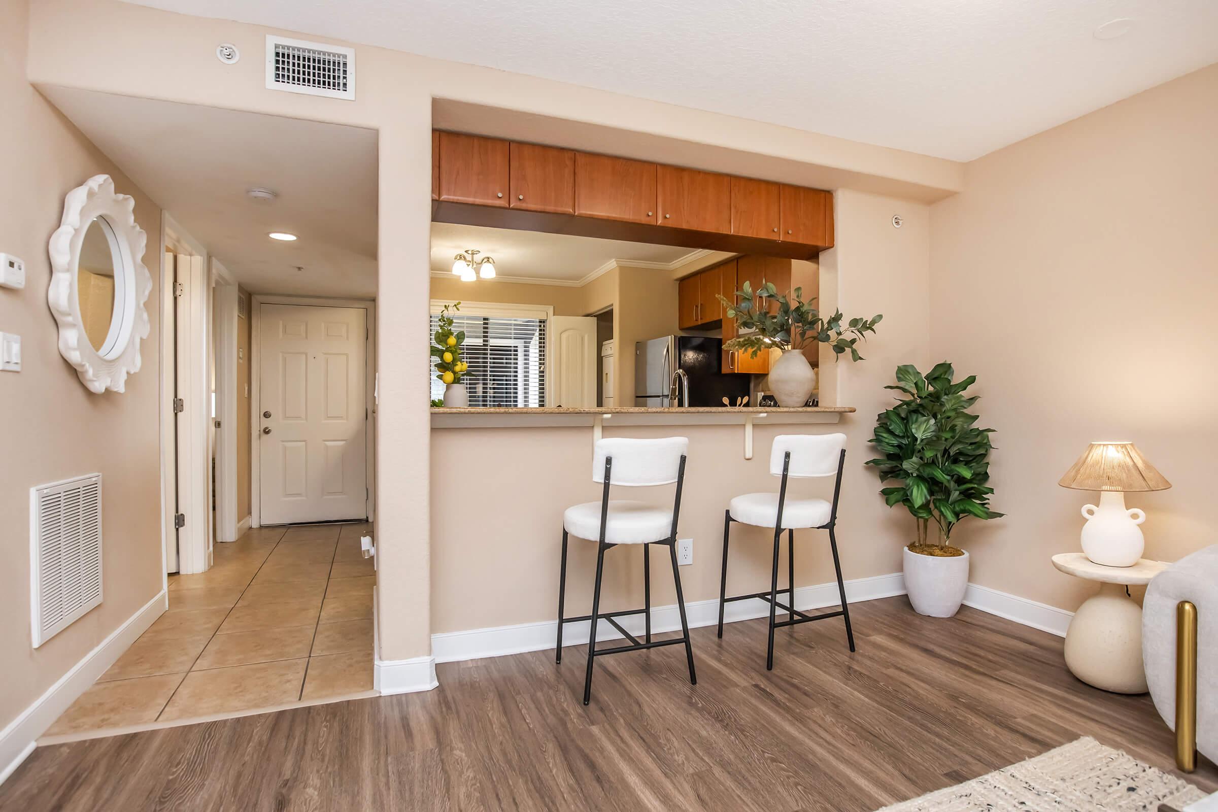A cozy interior view of a modern apartment kitchen and dining area. Features include a bar with two stools, a plant, a table lamp, and a doorway leading to a hallway. The warm colors and natural light create an inviting atmosphere.